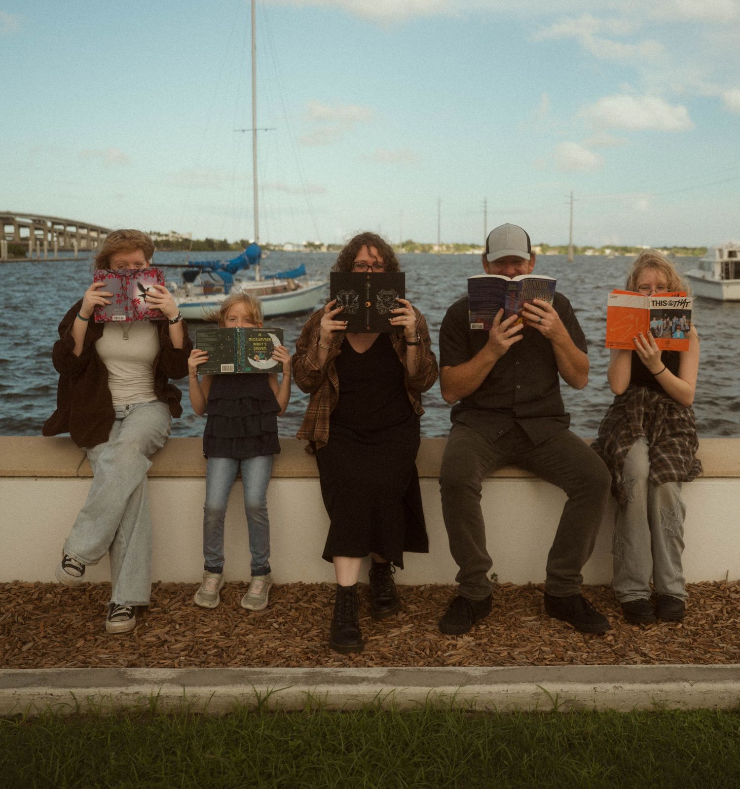 Five people sitting on a waterfront bench hiding their faces behind books, with sailboats and a bridge in the background.