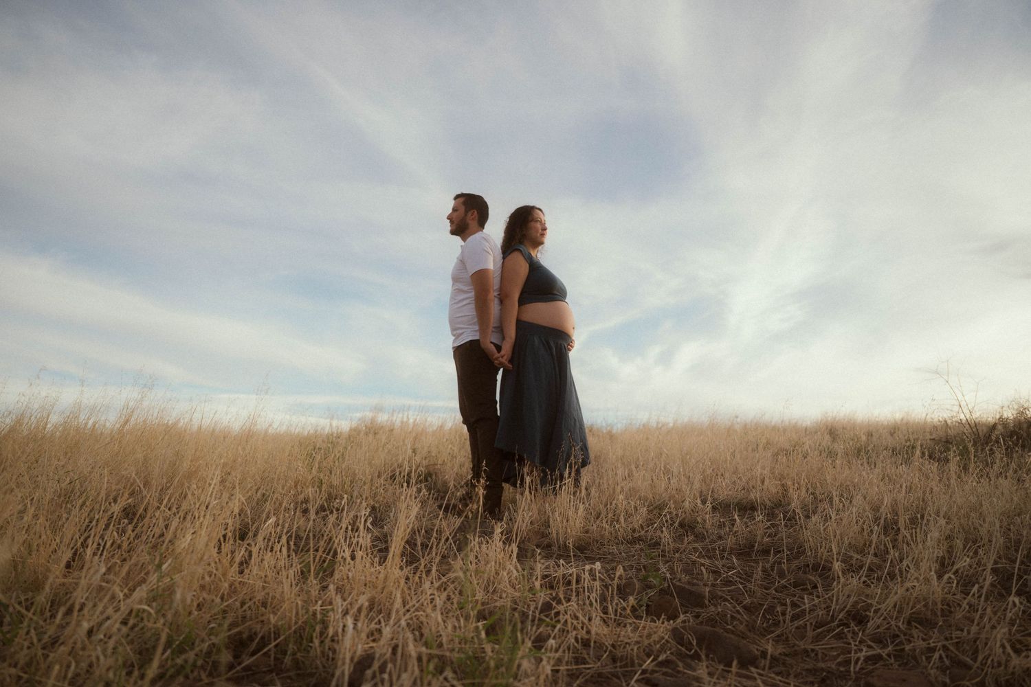 Pregnant couple standing back-to-back in golden field under cloudy sky during maternity photo shoot.