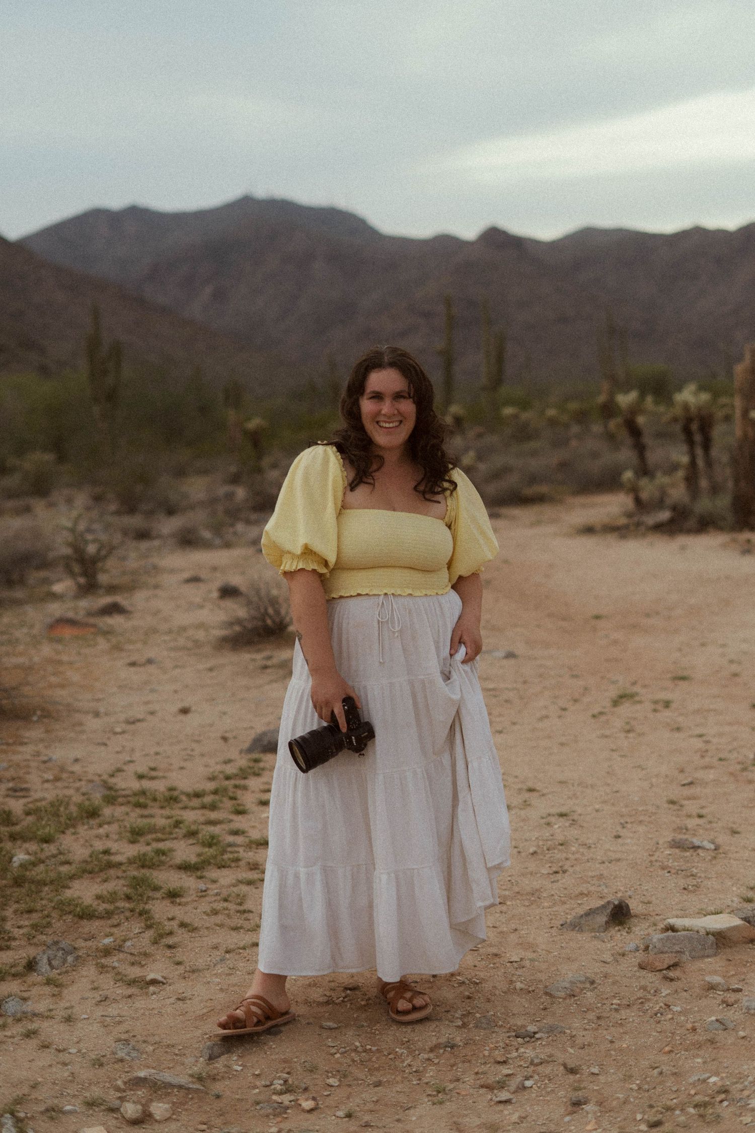 A woman in a yellow and white prairie dress stands in a desert landscape with mountains and saguaro cacti in the background.