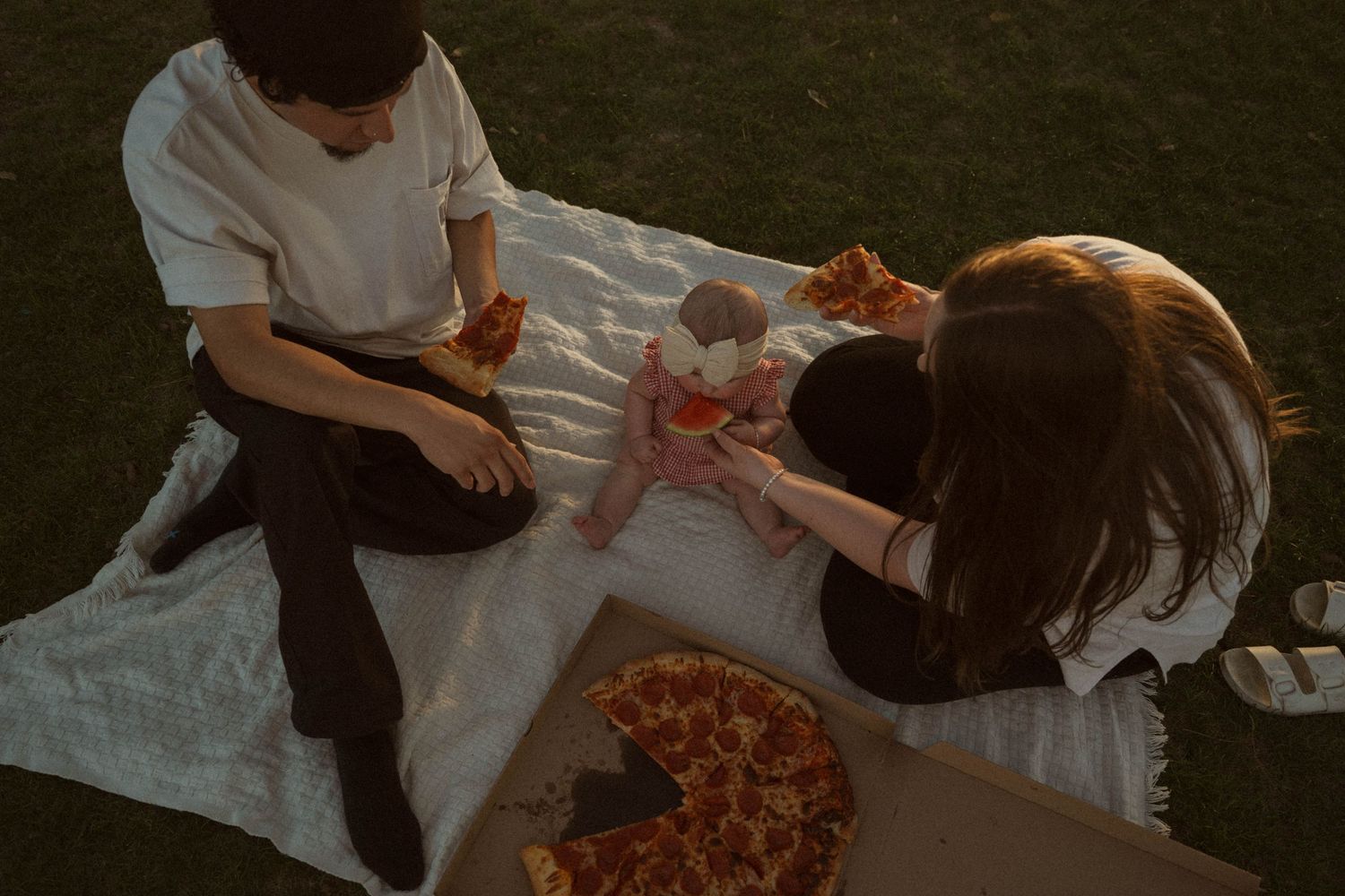 Two people enjoying a pizza picnic on a white blanket in a park during golden hour sunset.