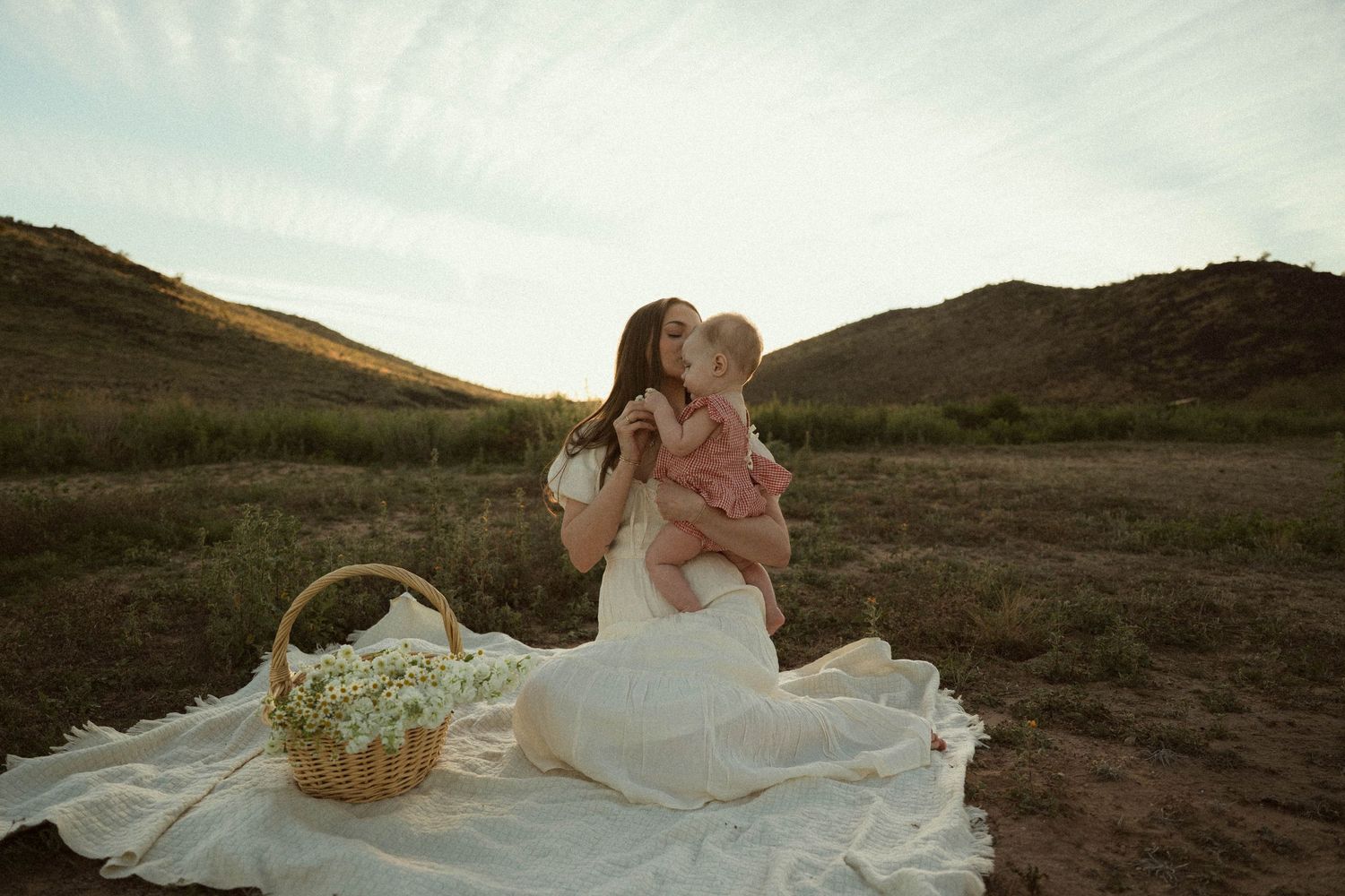 A woman in a white dress sits on a blanket outdoors, kissing a baby, with a wicker basket nearby and hills in the background.