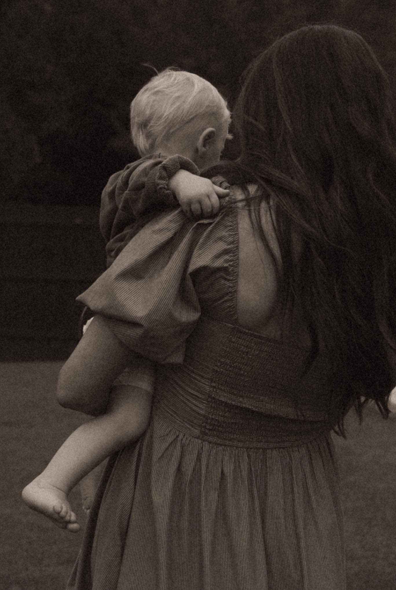Black and white photo of a mother with long dark hair holding a blonde toddler in a dress outdoors.