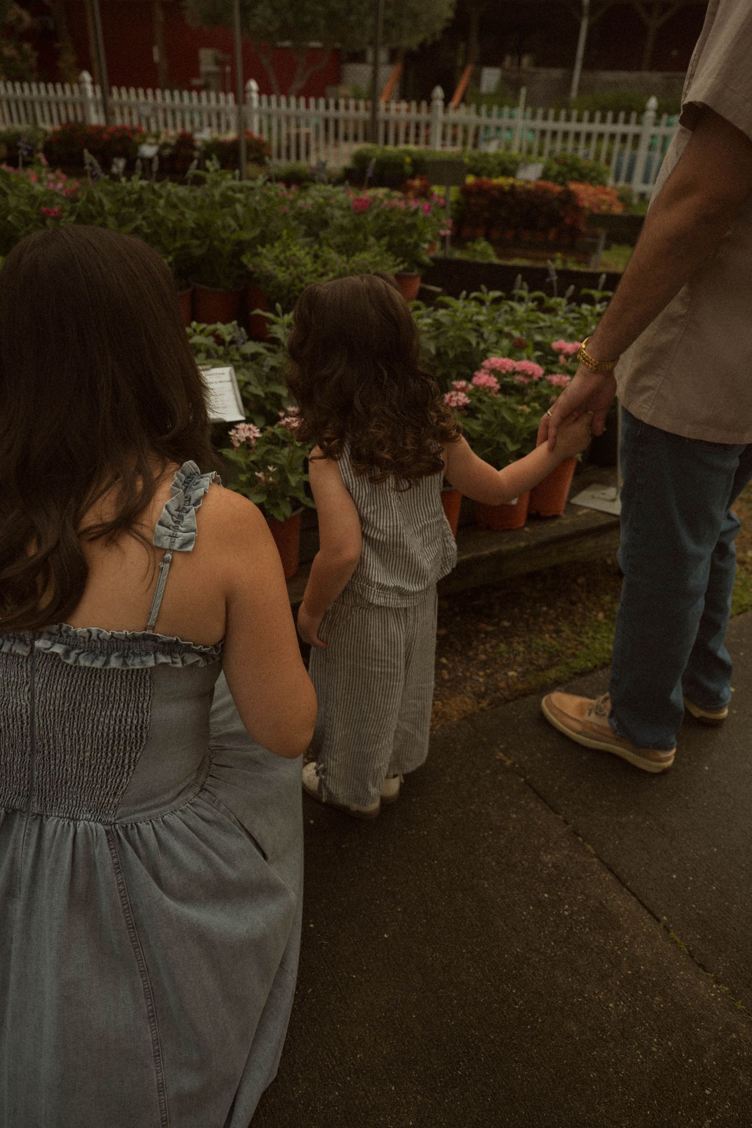 Two girls and an adult explore a garden nursery filled with colorful flowers and green plants on a cloudy day.