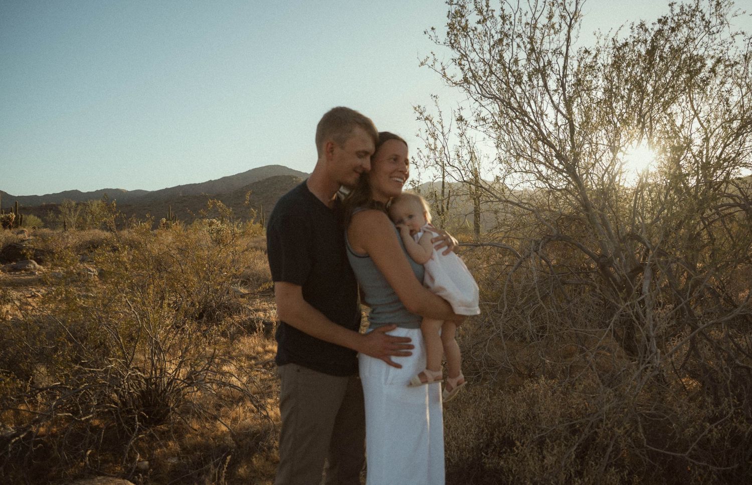 Couple holding baby in desert landscape at golden hour with mountains and cacti in background.