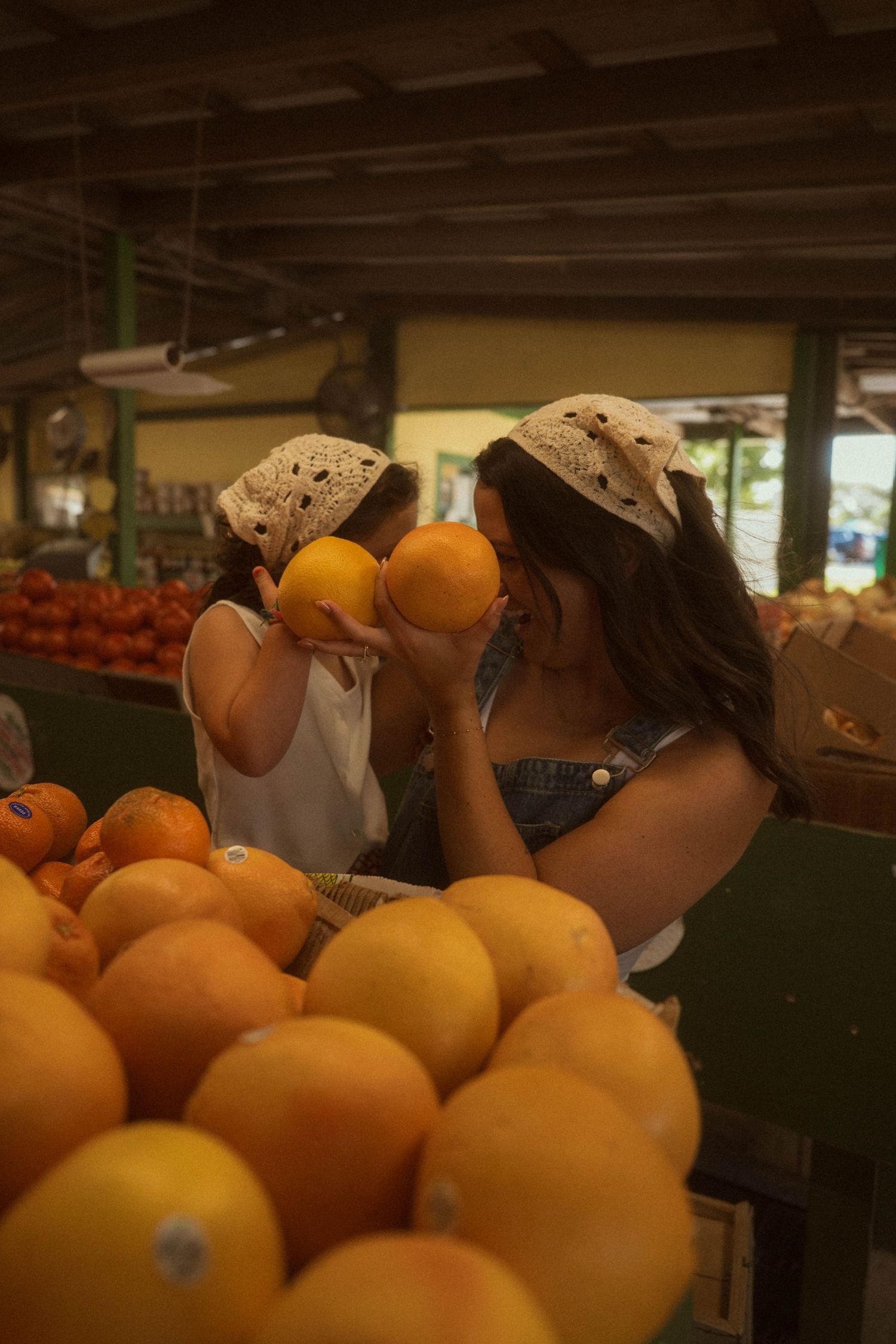 Two women wearing matching bandanas smell fresh oranges at a vibrant outdoor fruit market.
