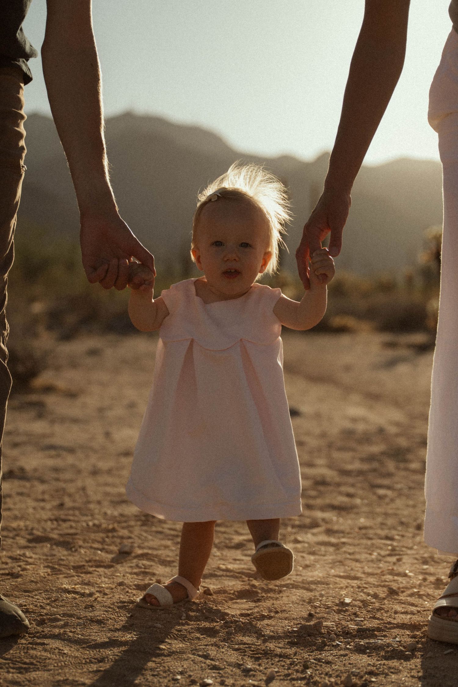 A toddler in a pink dress walks hand-in-hand with parents on a desert path with mountains in the background.