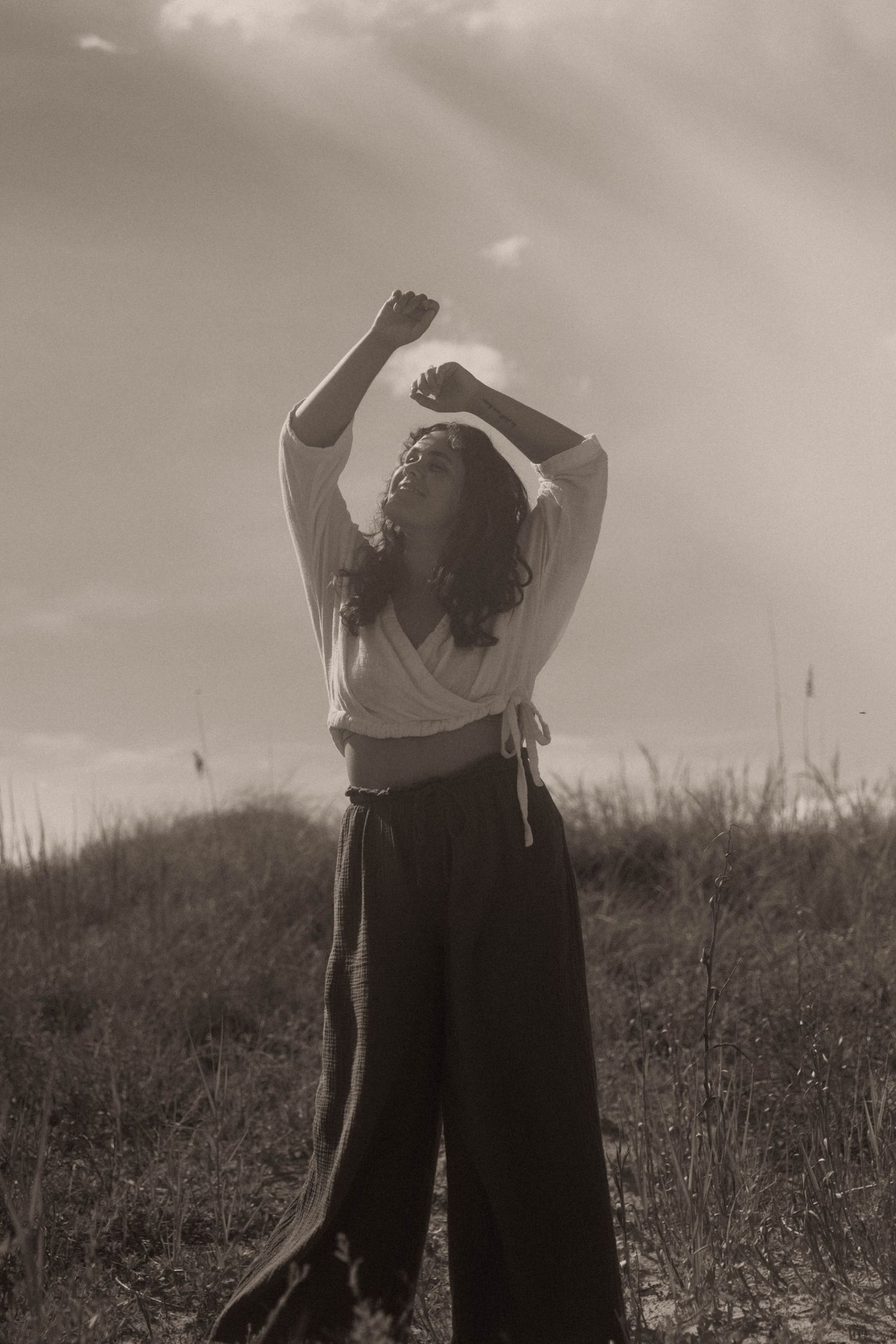 A woman in wide-leg pants raises her arms toward the sky in a serene sepia-toned outdoor portrait.