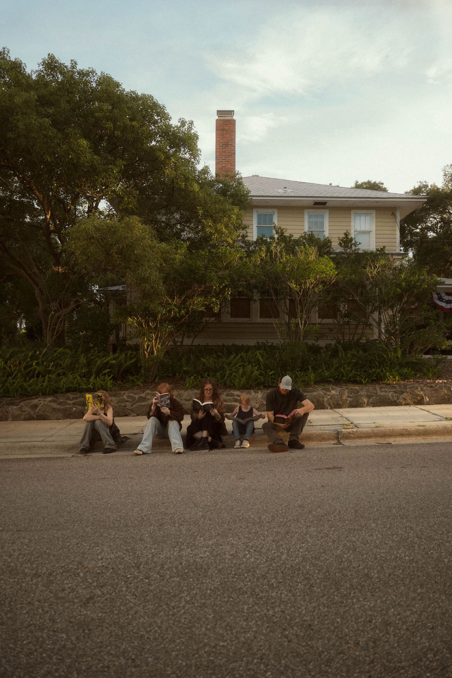 Group of friends sitting on a curb reading books in front of a house with trees and a brick chimney.