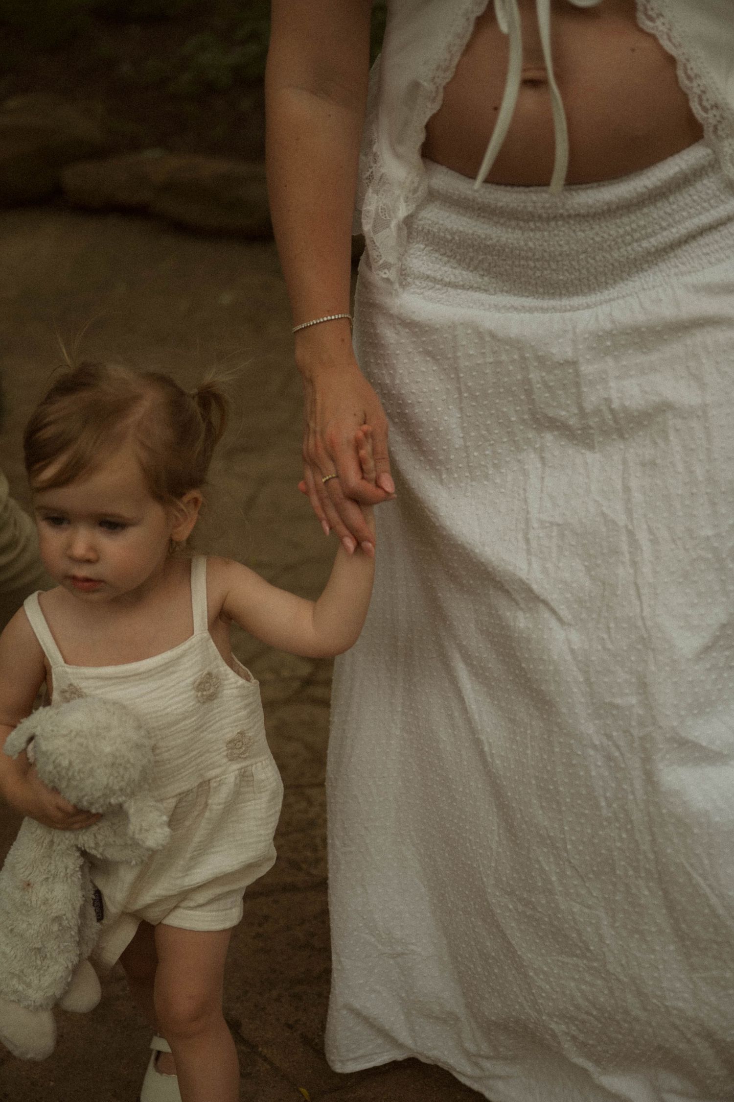 Pregnant woman in white dress holding hands with toddler girl carrying stuffed animal toy outdoors.