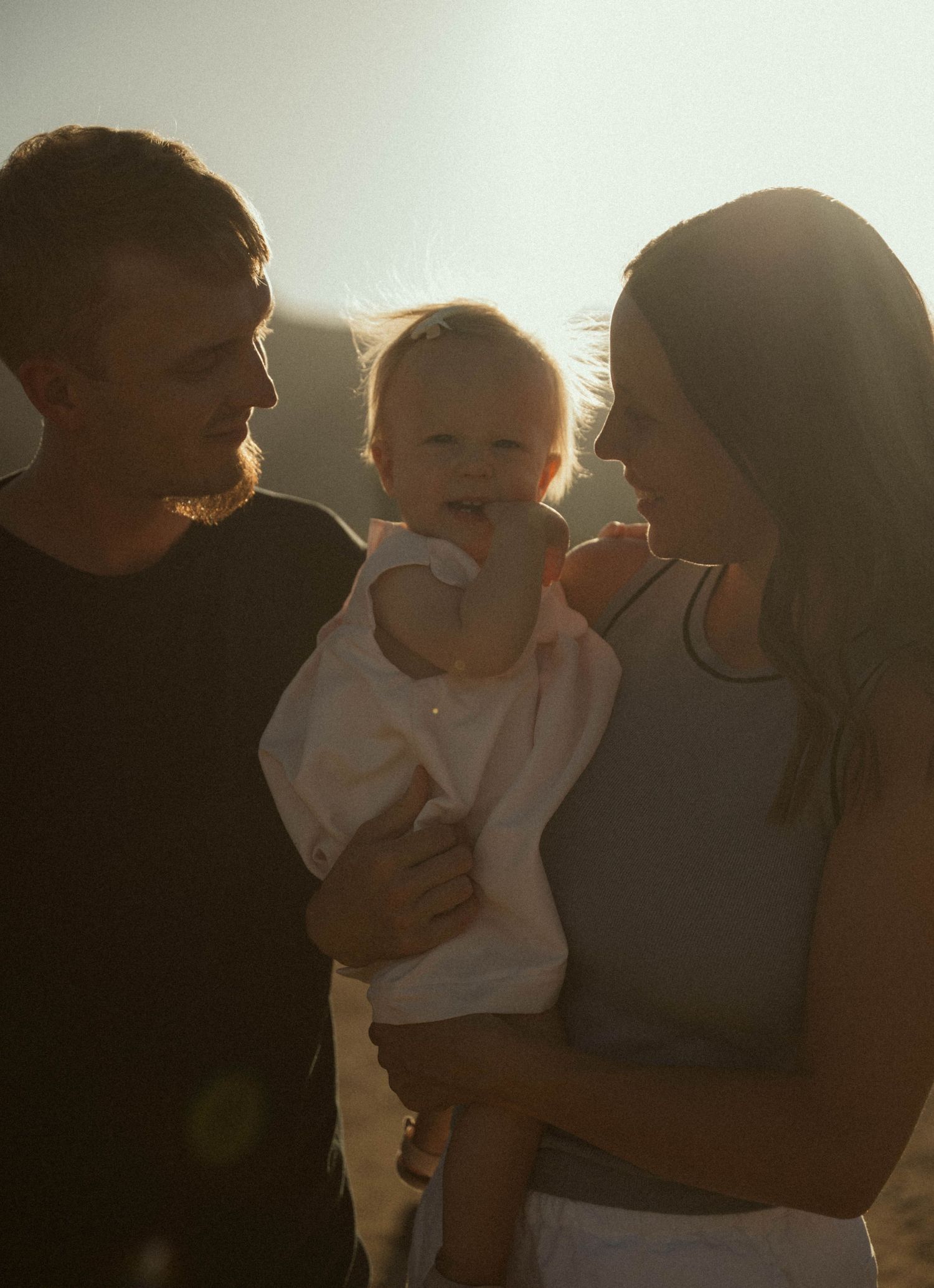 A young couple smiles at their baby in warm golden backlit sunlight outdoors.