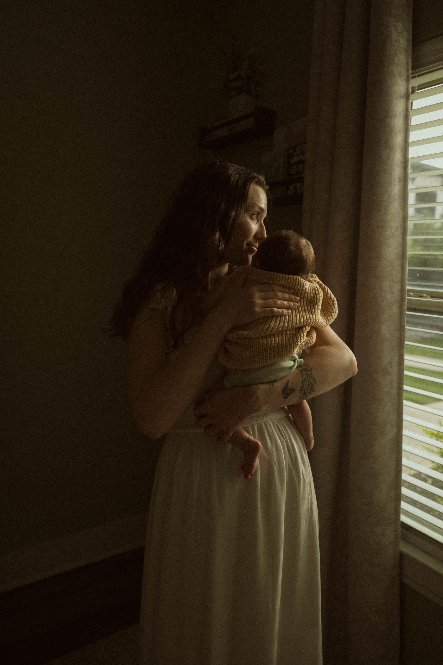 A young mother in a white dress holds her newborn baby near a window with soft natural light.