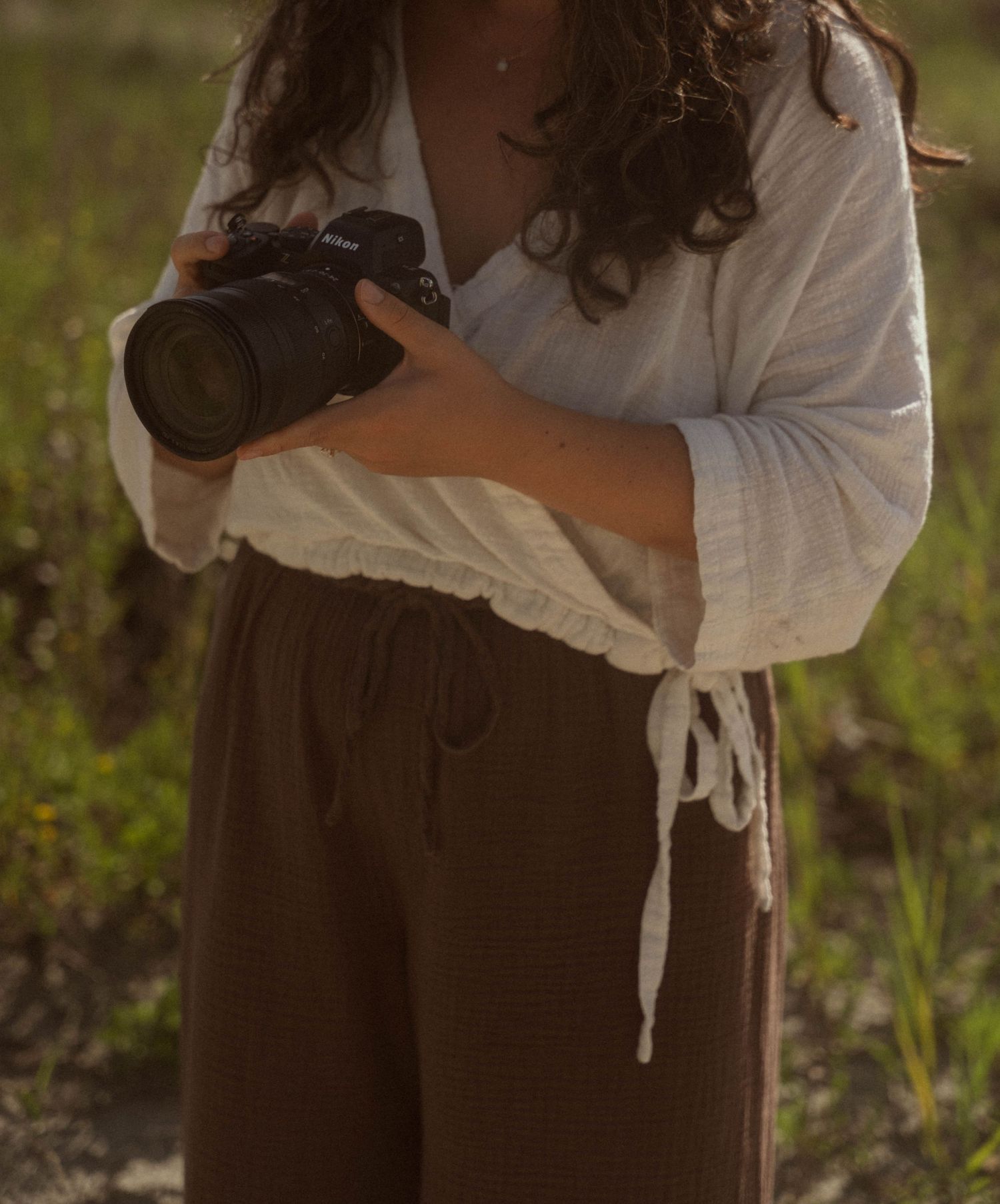 Woman in white linen top and brown pants holding a Canon camera outdoors in a natural setting.