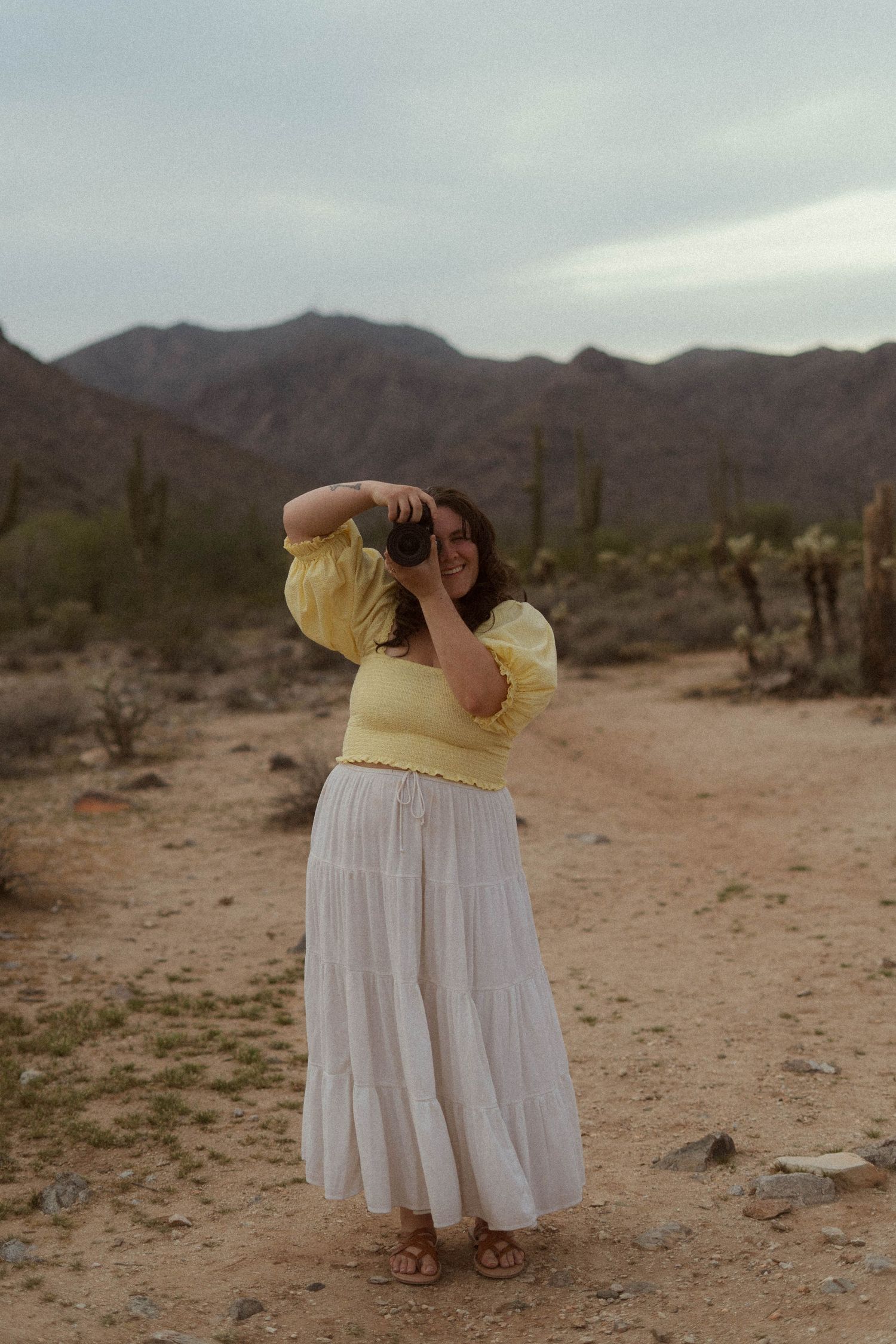 Two figures in white dresses embrace against a desert landscape with mountains and saguaro cacti in the background.