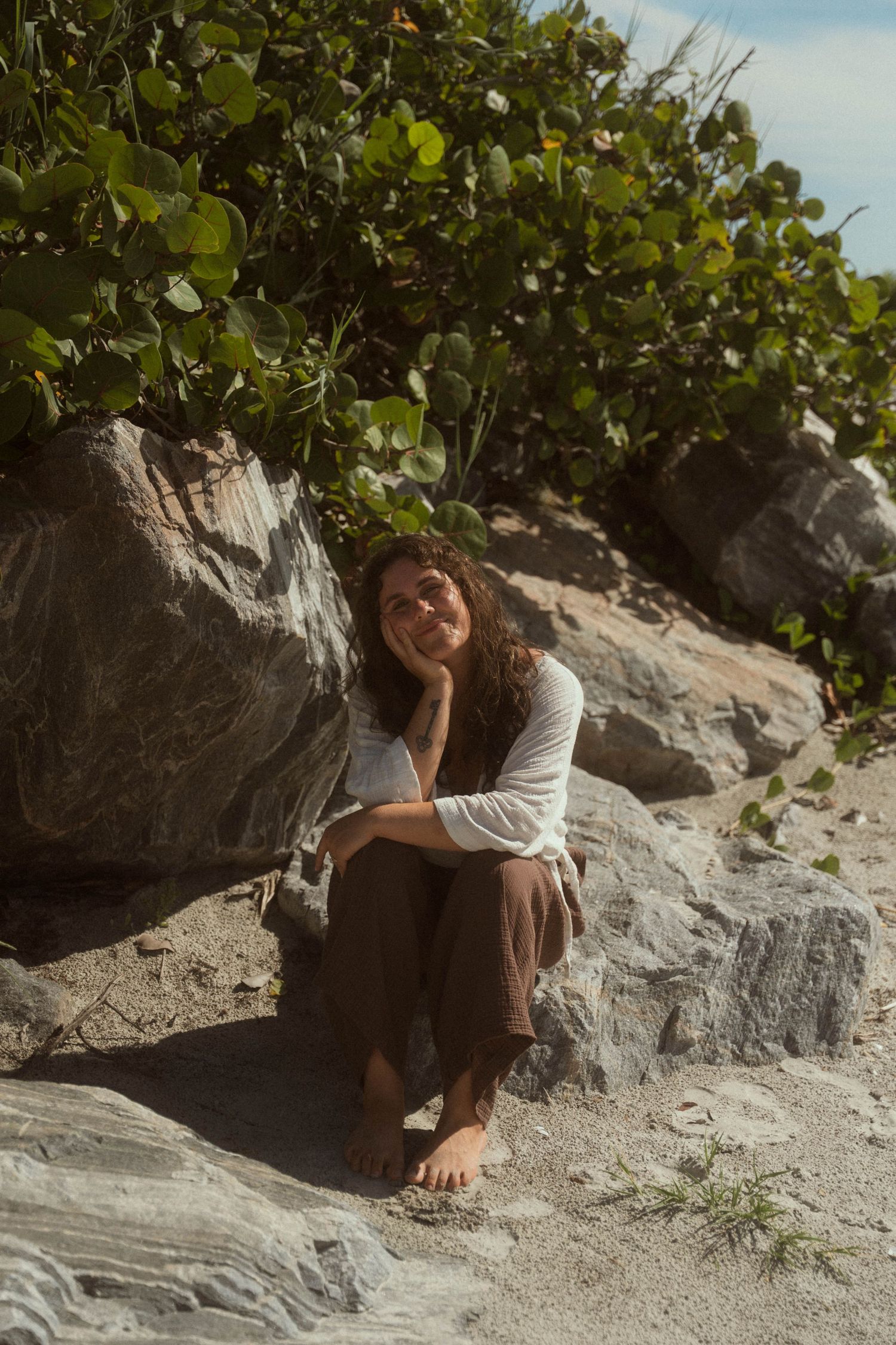 Woman sitting barefoot on rocky beach, leaning against boulder, smiling upward, surrounded by lush green foliage.