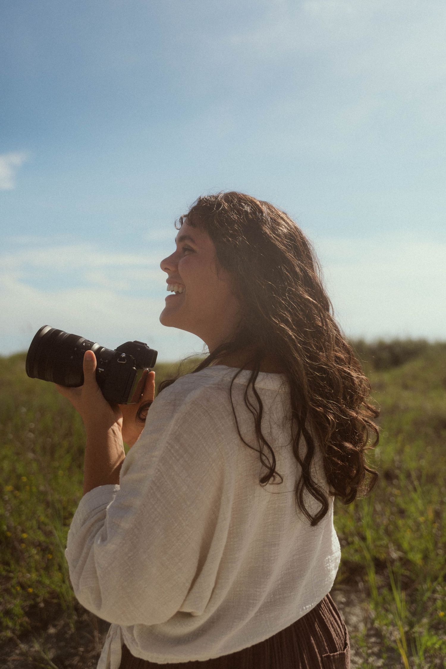 Smiling woman with curly hair holding a camera outdoors in a sunny green field wearing a white linen top.
