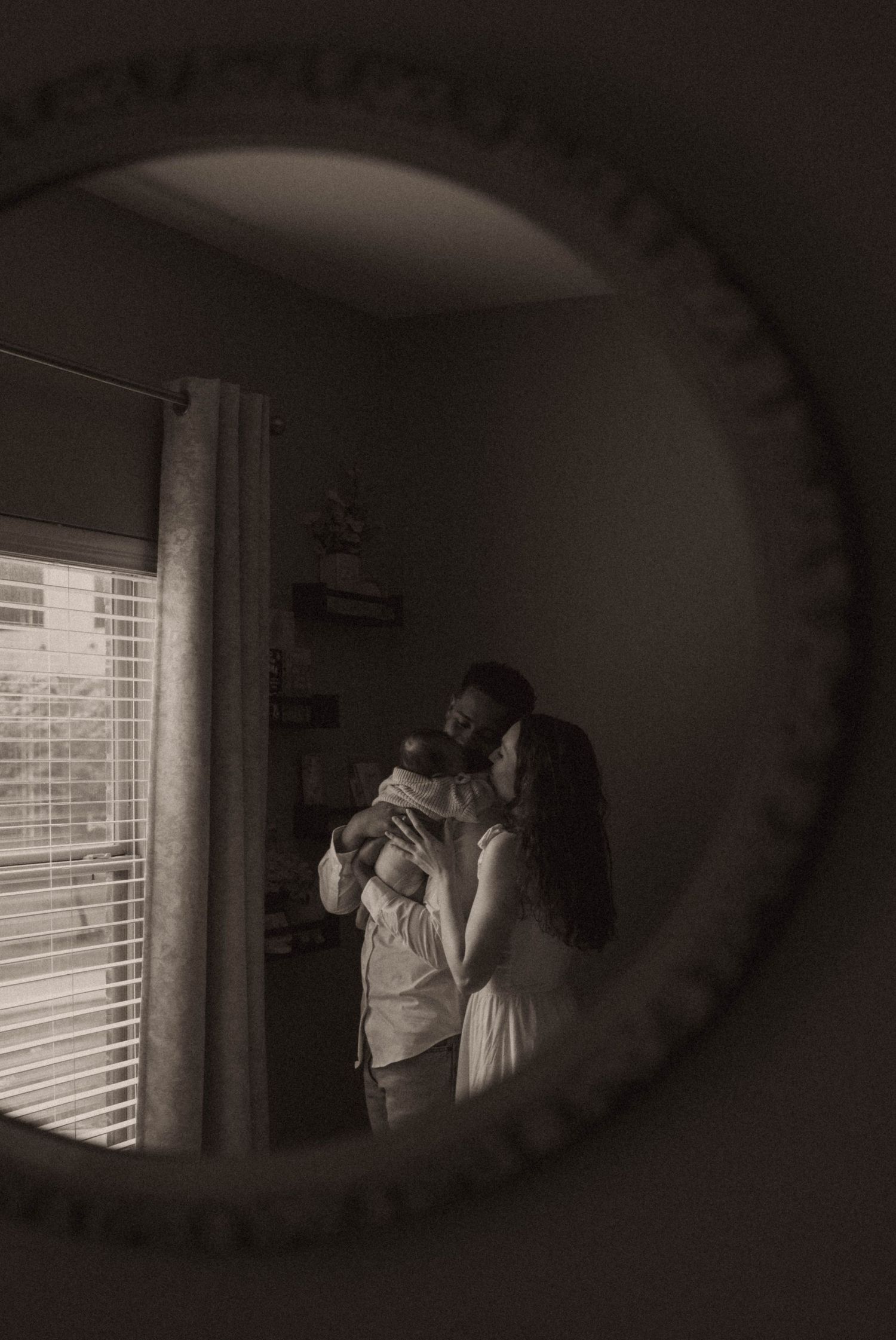 Couple sharing a tender moment holding newborn baby, reflected in ornate round mirror, black and white photo.