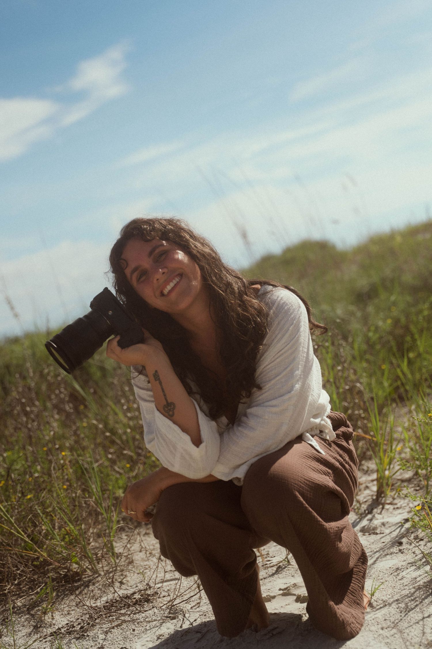 Smiling woman crouching outdoors holding a camera, with sandy dunes and blue sky in the background.