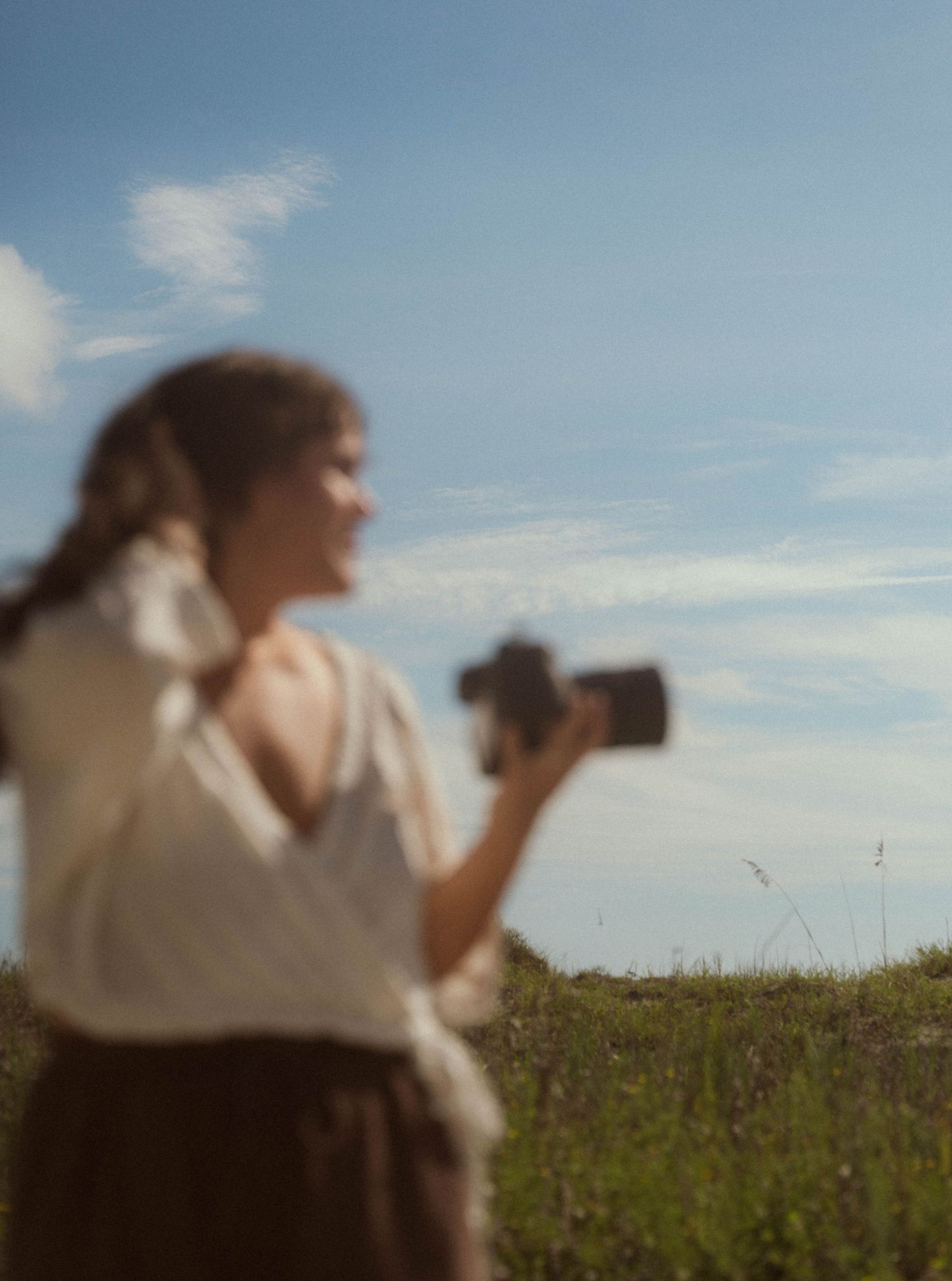 A blurred woman holds a camera in a sunny green field under a blue sky.