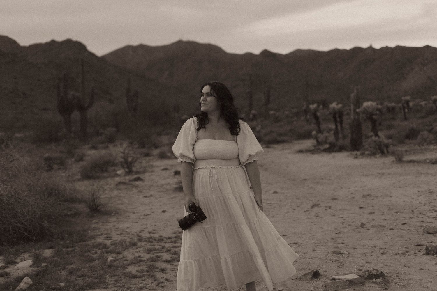 A person in a white flowing dress poses dramatically against a desert landscape at sunset.