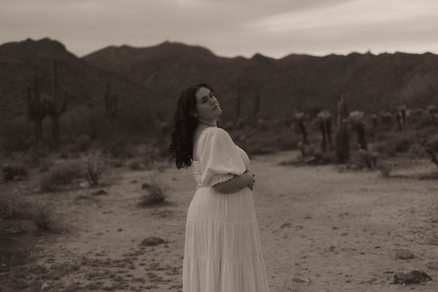 A figure in a flowing white dress poses gracefully in a desert landscape with mountains and cacti in the background.