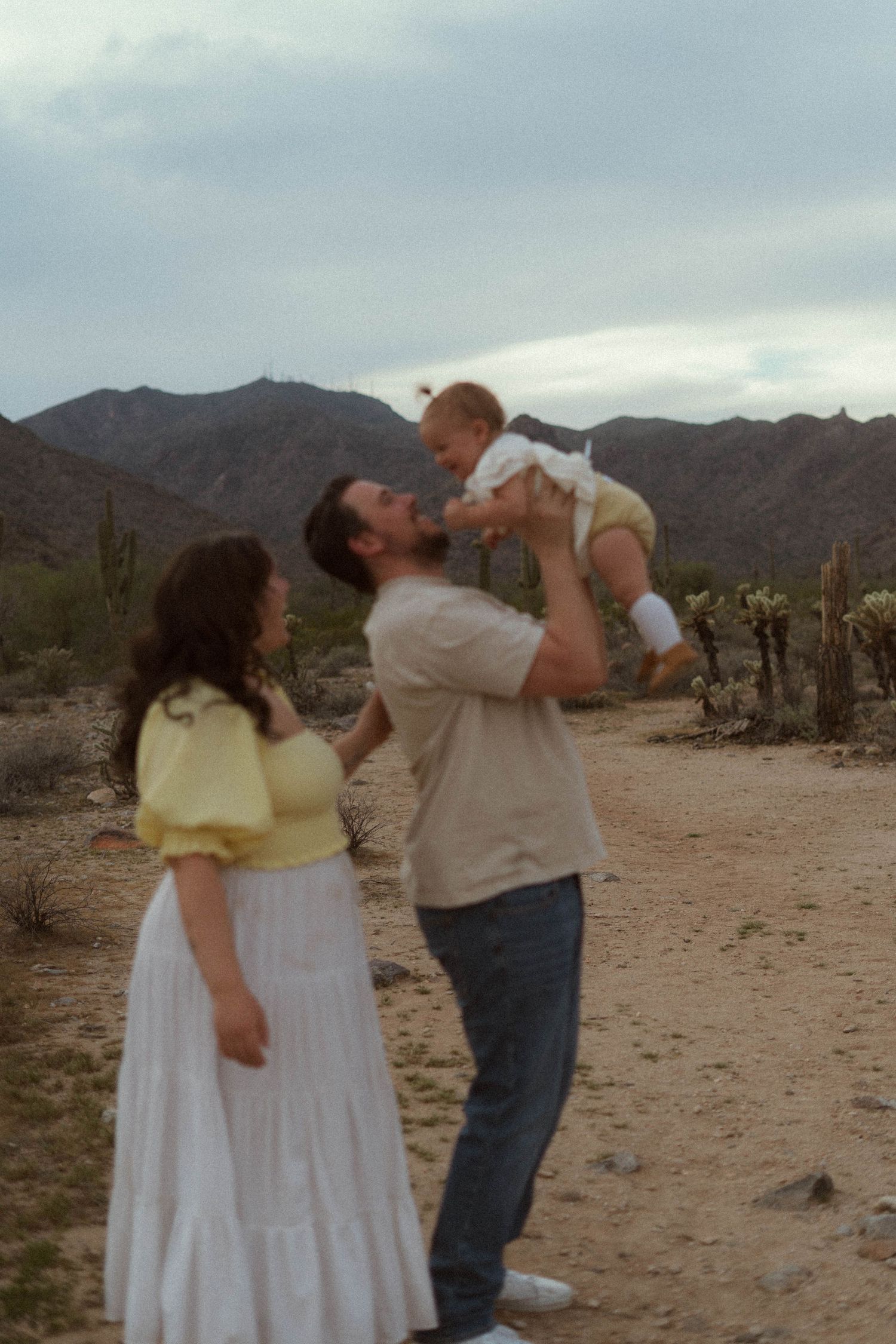 A couple in desert attire shares a drink from water bottles in a scenic desert landscape with mountains in the background.