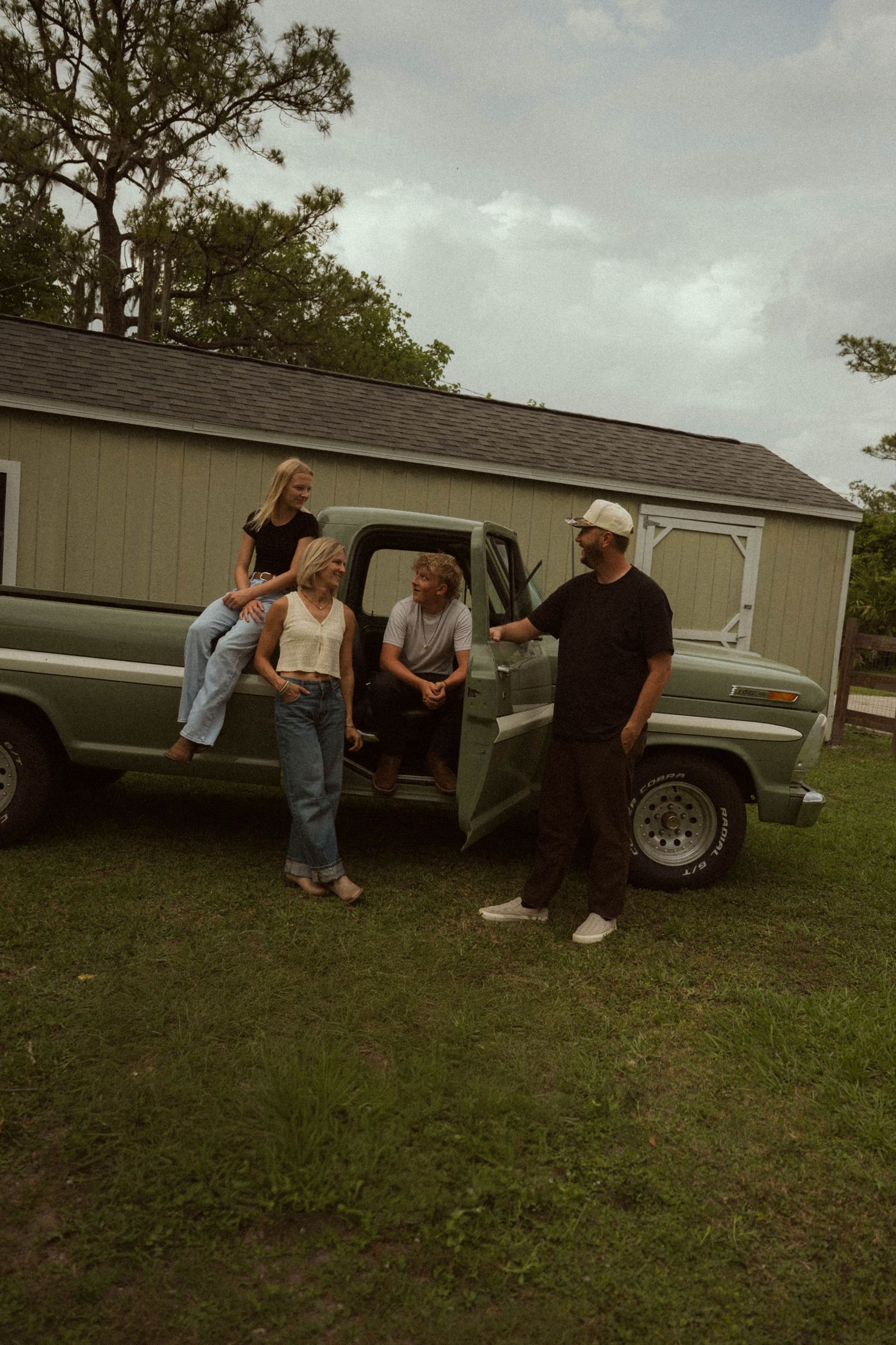 Four people gathered around a vintage green pickup truck on a grassy lawn beside a gray shed.