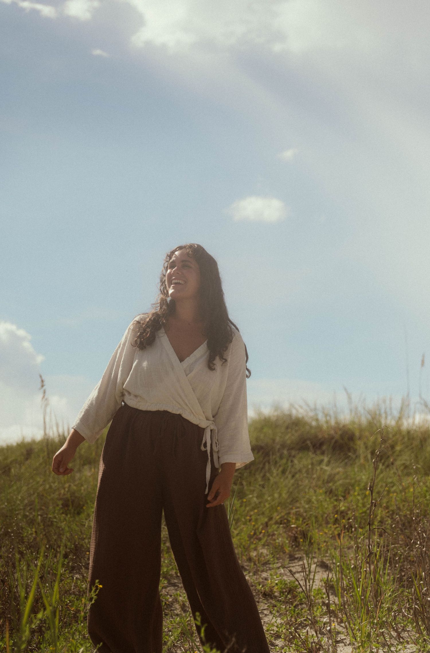 A woman in a white wrap top and dark wide-leg pants stands on a grassy dune under a bright sky.