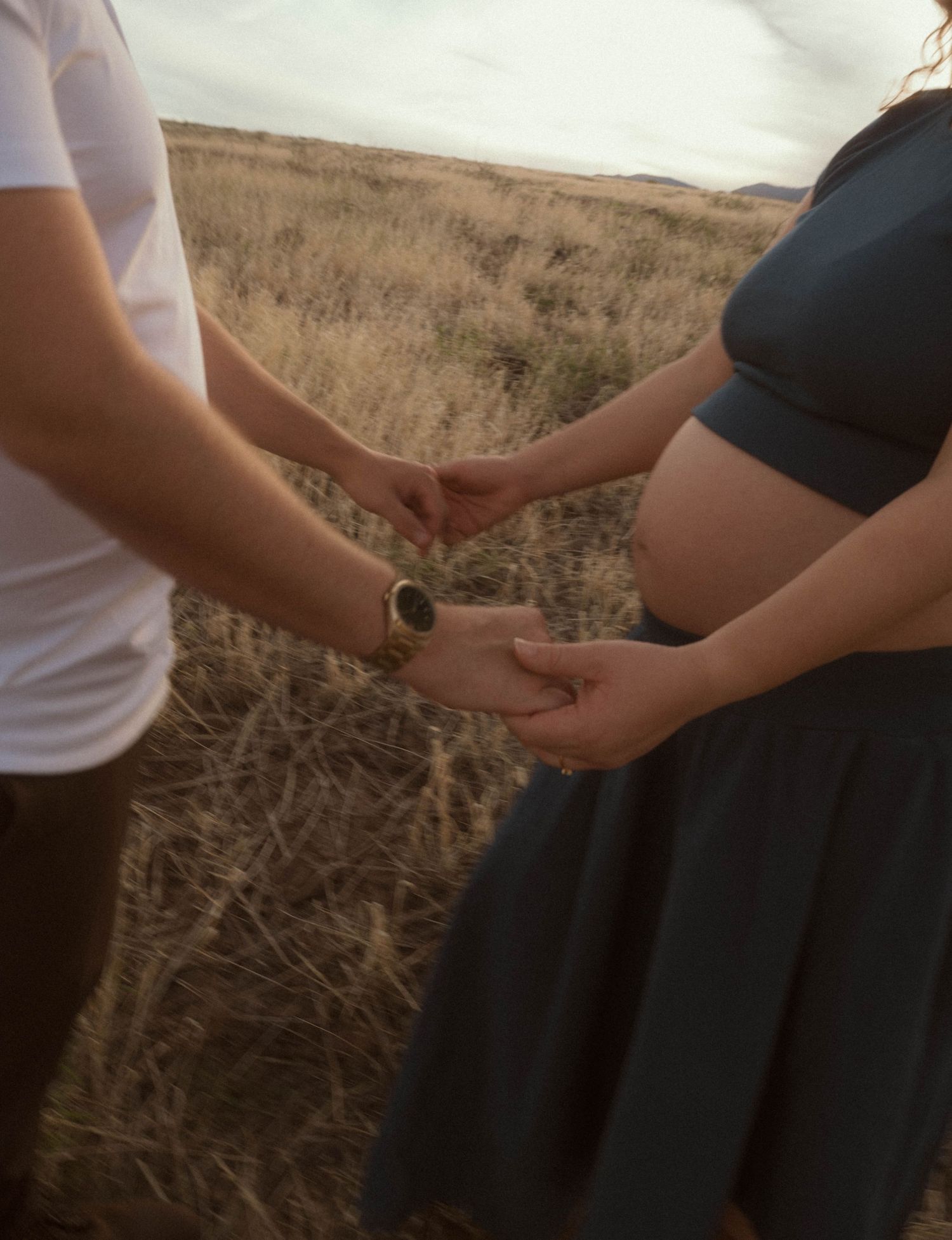 A pregnant couple holds hands in a golden field, showcasing a maternity photo shoot outdoors.