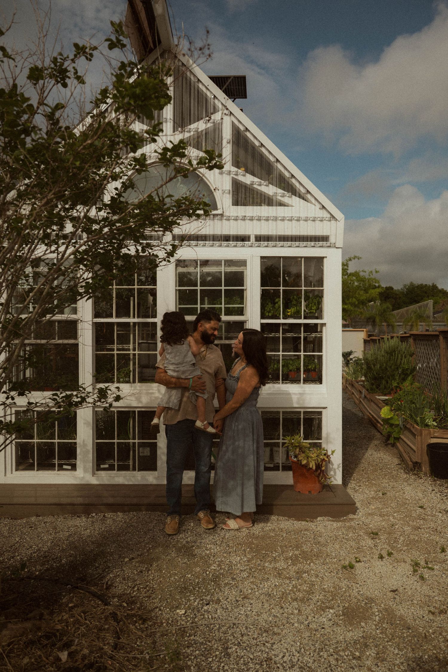 A family of three poses in front of a white greenhouse surrounded by lush greenery and potted plants.