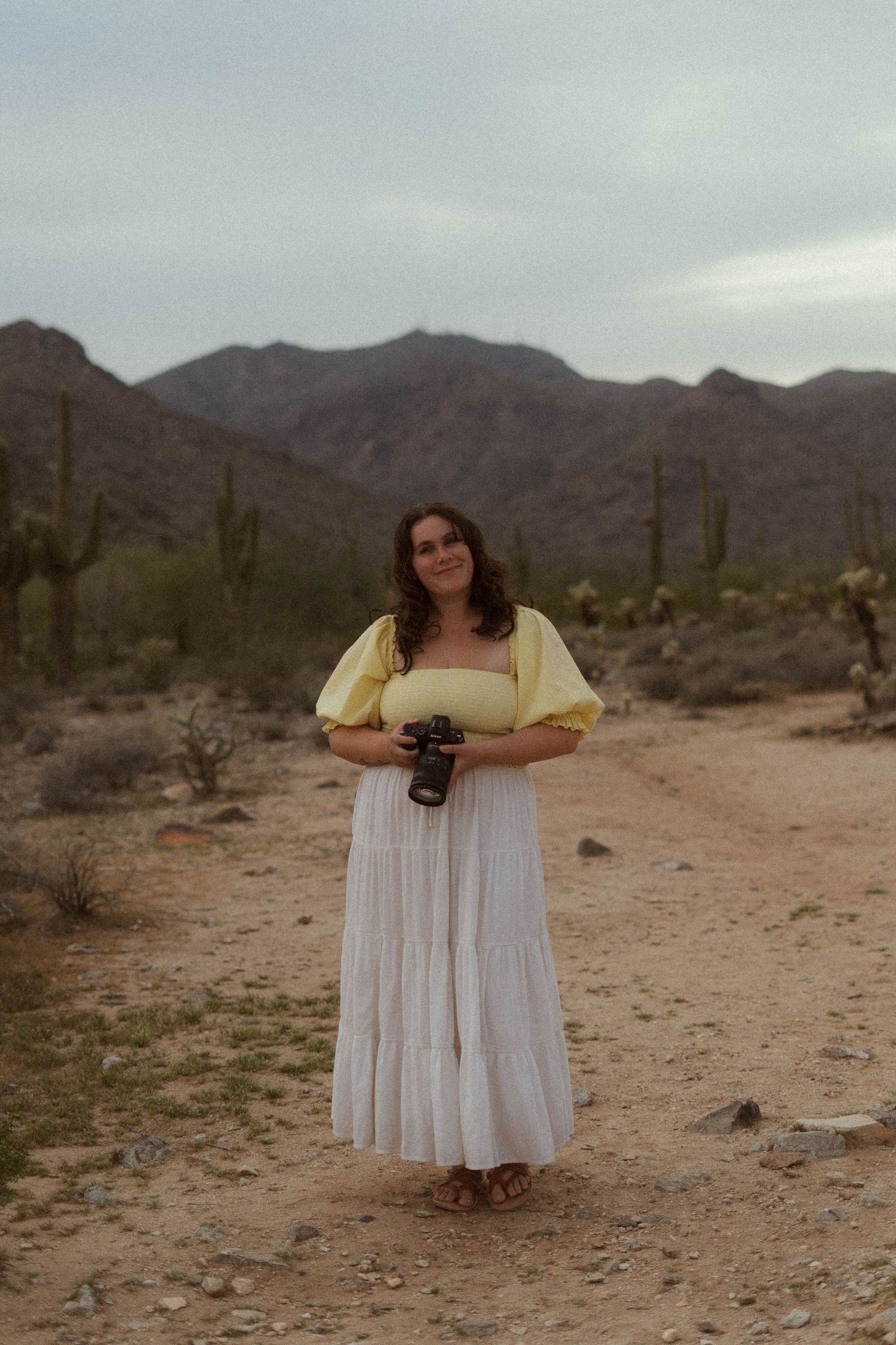 A person in a white flowing dress poses in a desert landscape with mountains and saguaro cacti in the background.