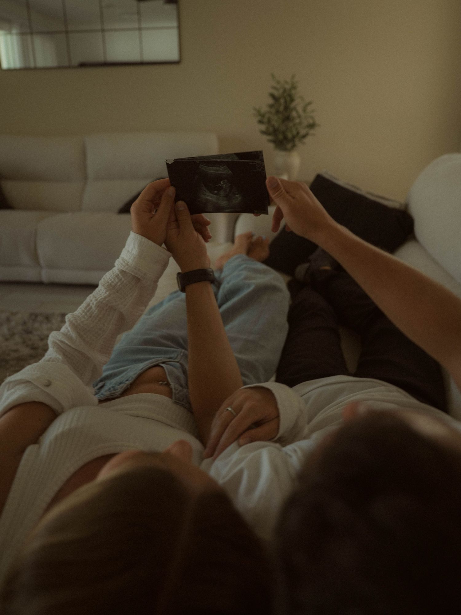 Couple lying together holding up an ultrasound photo in a cozy living room, sharing a tender pregnancy announcement moment.