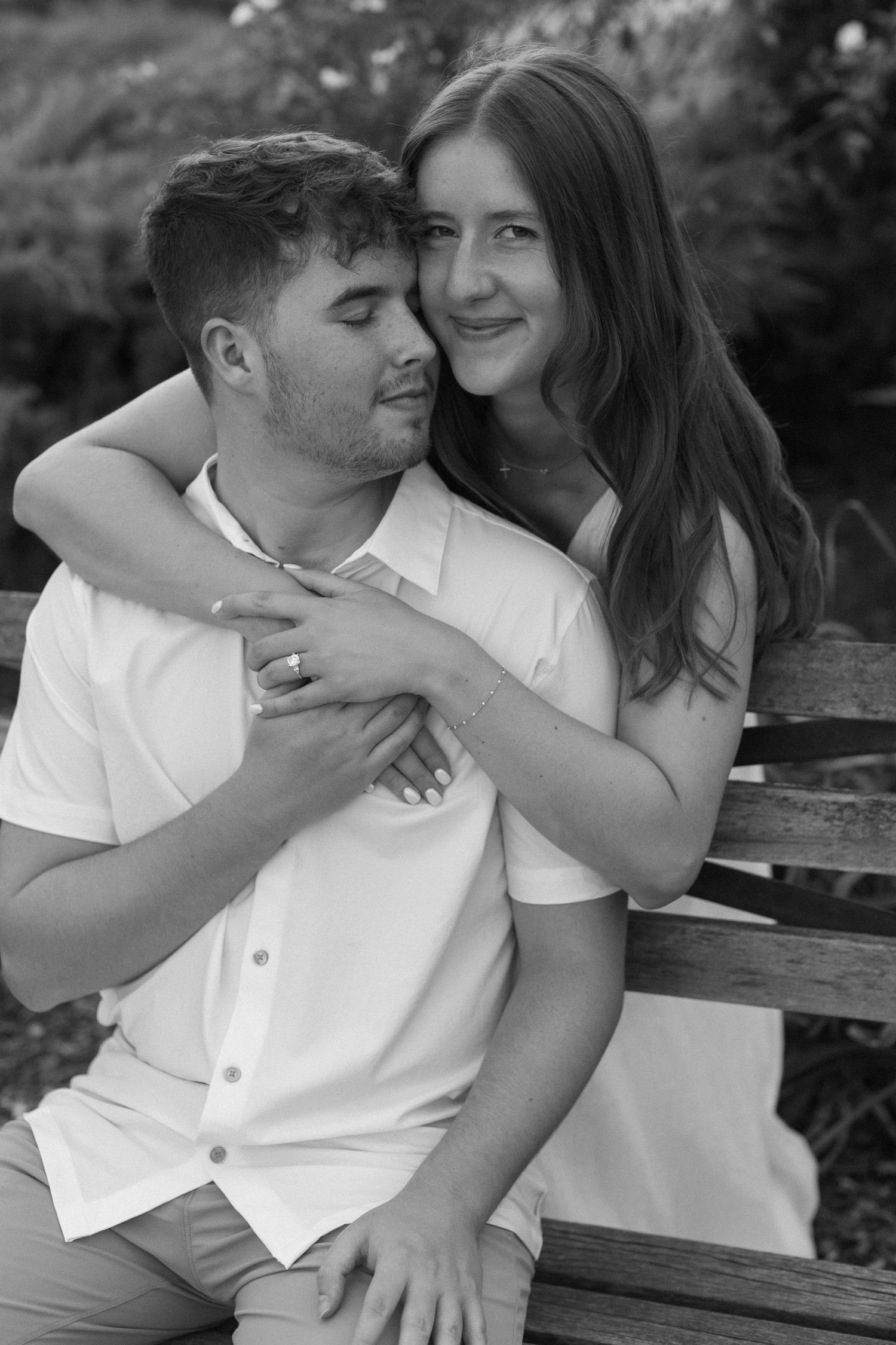 A romantic black and white photo of a couple embracing on a wooden bench in a garden setting.