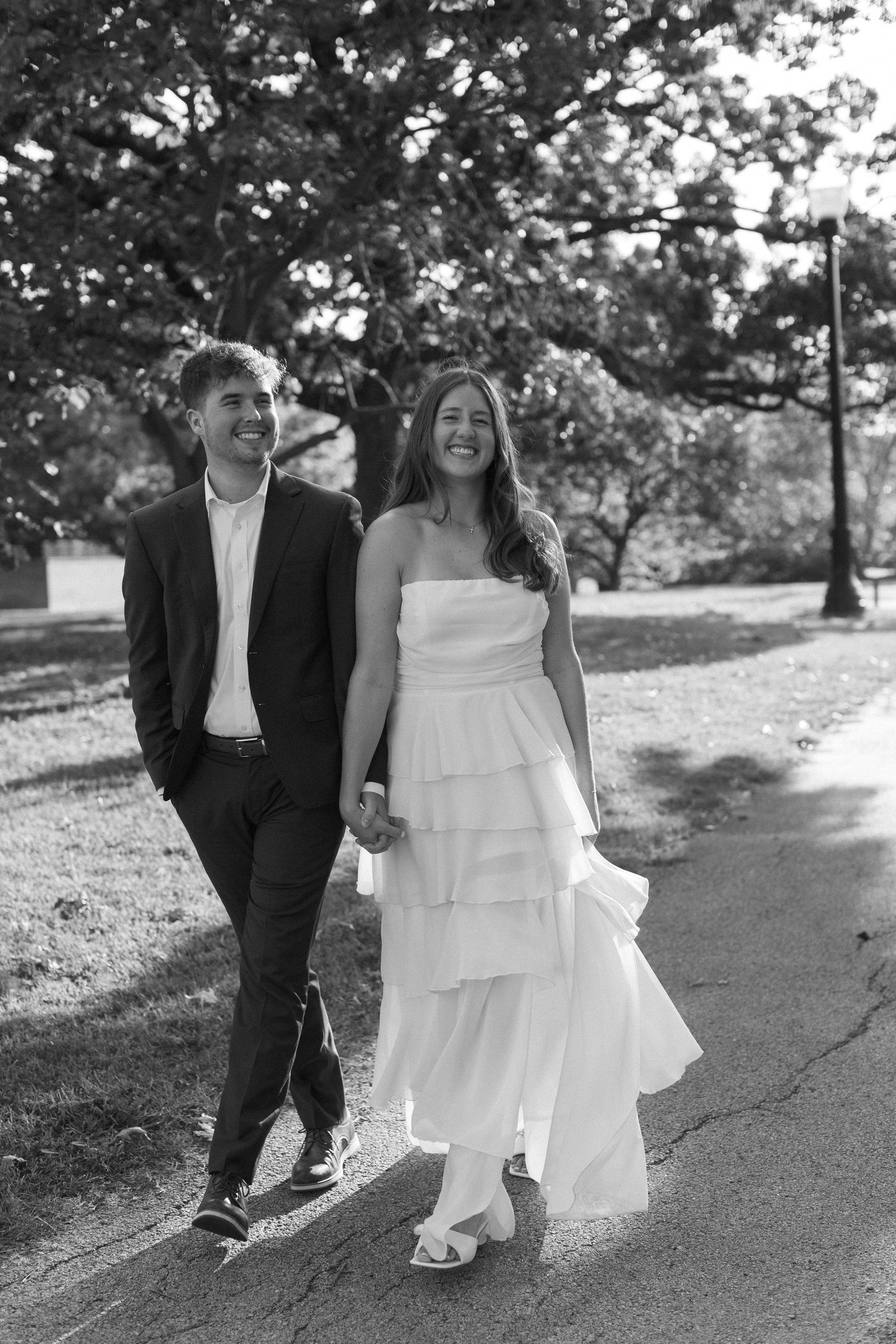 A romantic couple strolls down a park pathway in formal black and white outfits.