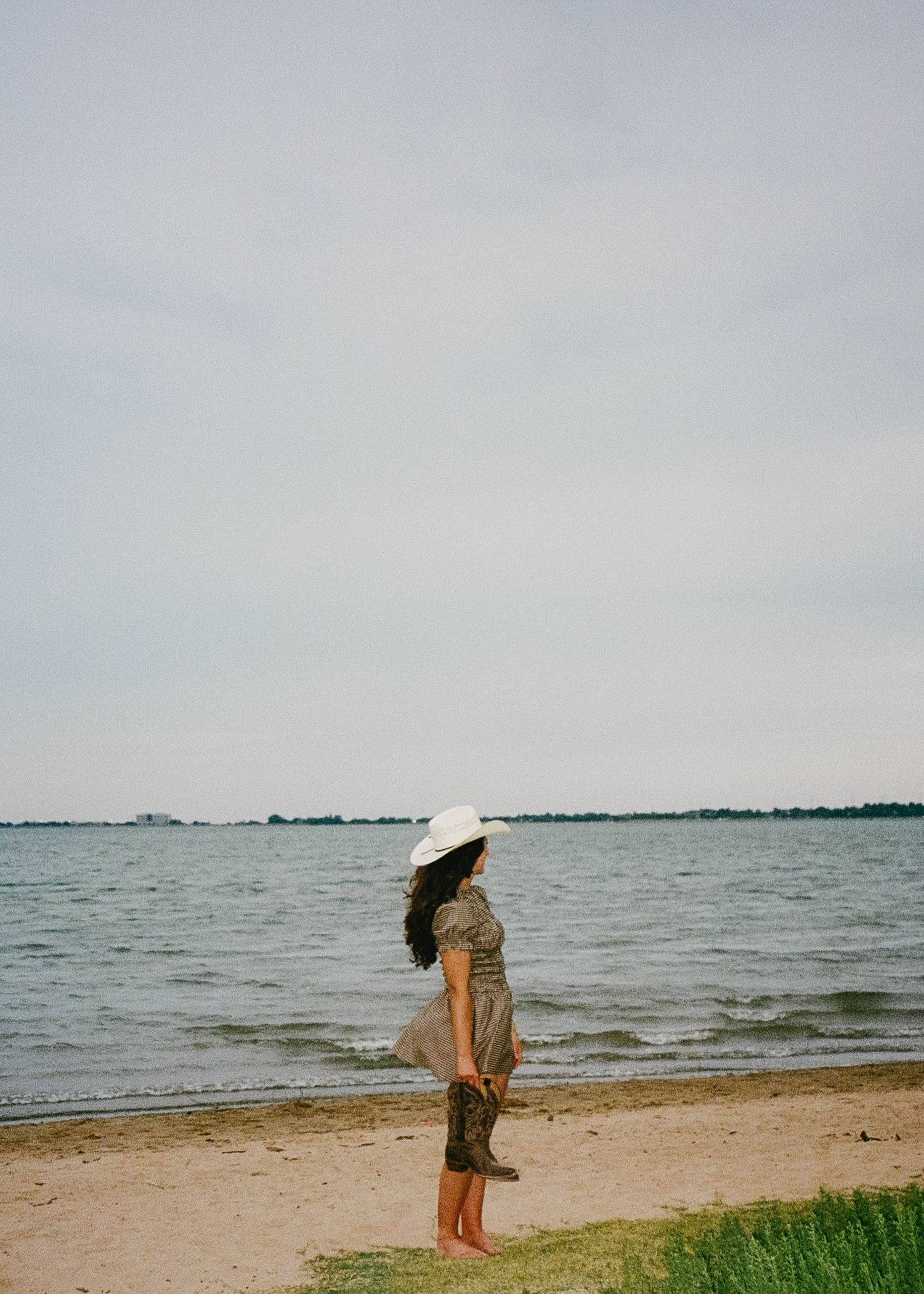 Someone in a sunhat and flowy dress stands contemplatively at the edge of a beach on an overcast day by the sea.