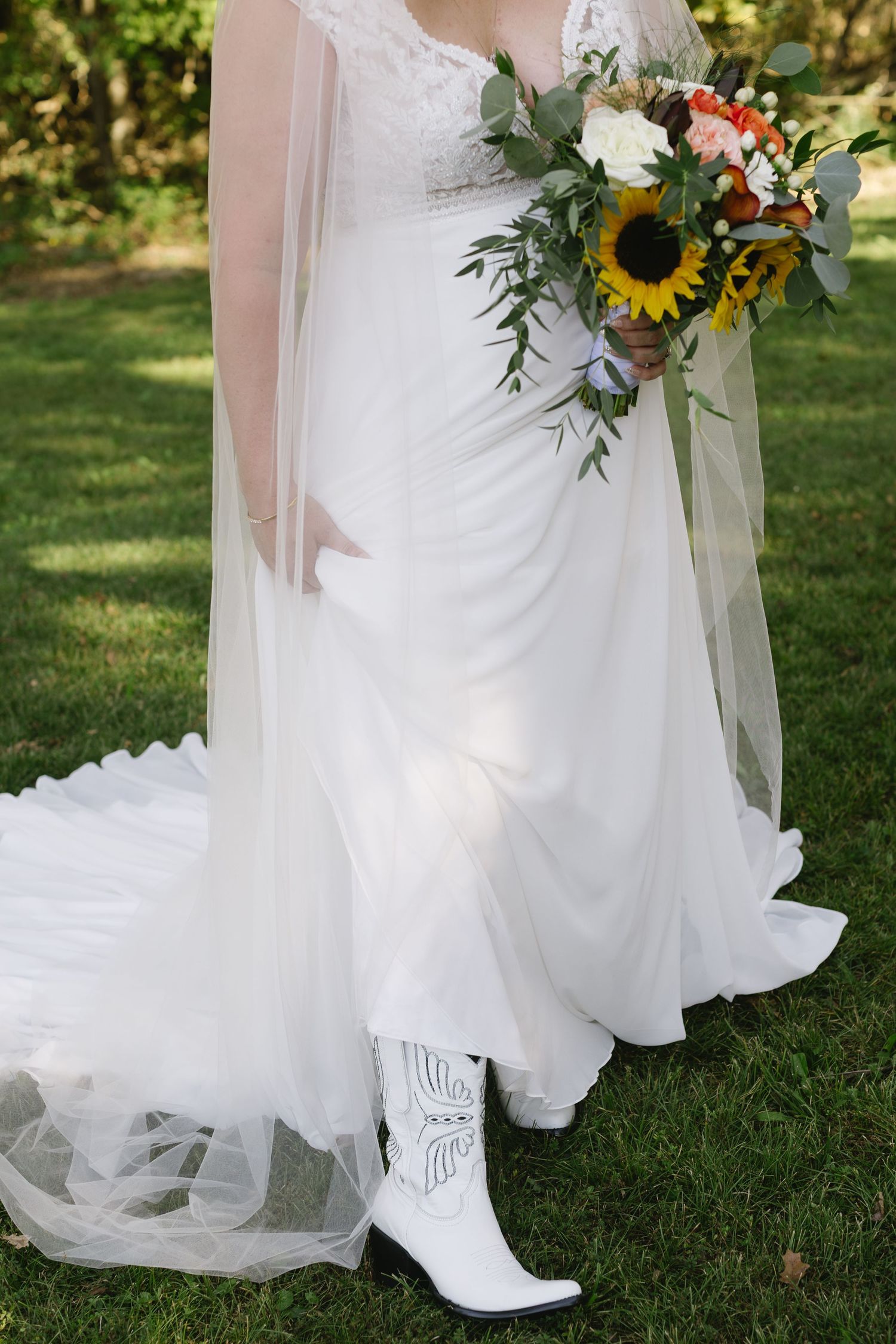White wedding dress with cowboy boots and sunflower bouquet details shown against green grass.