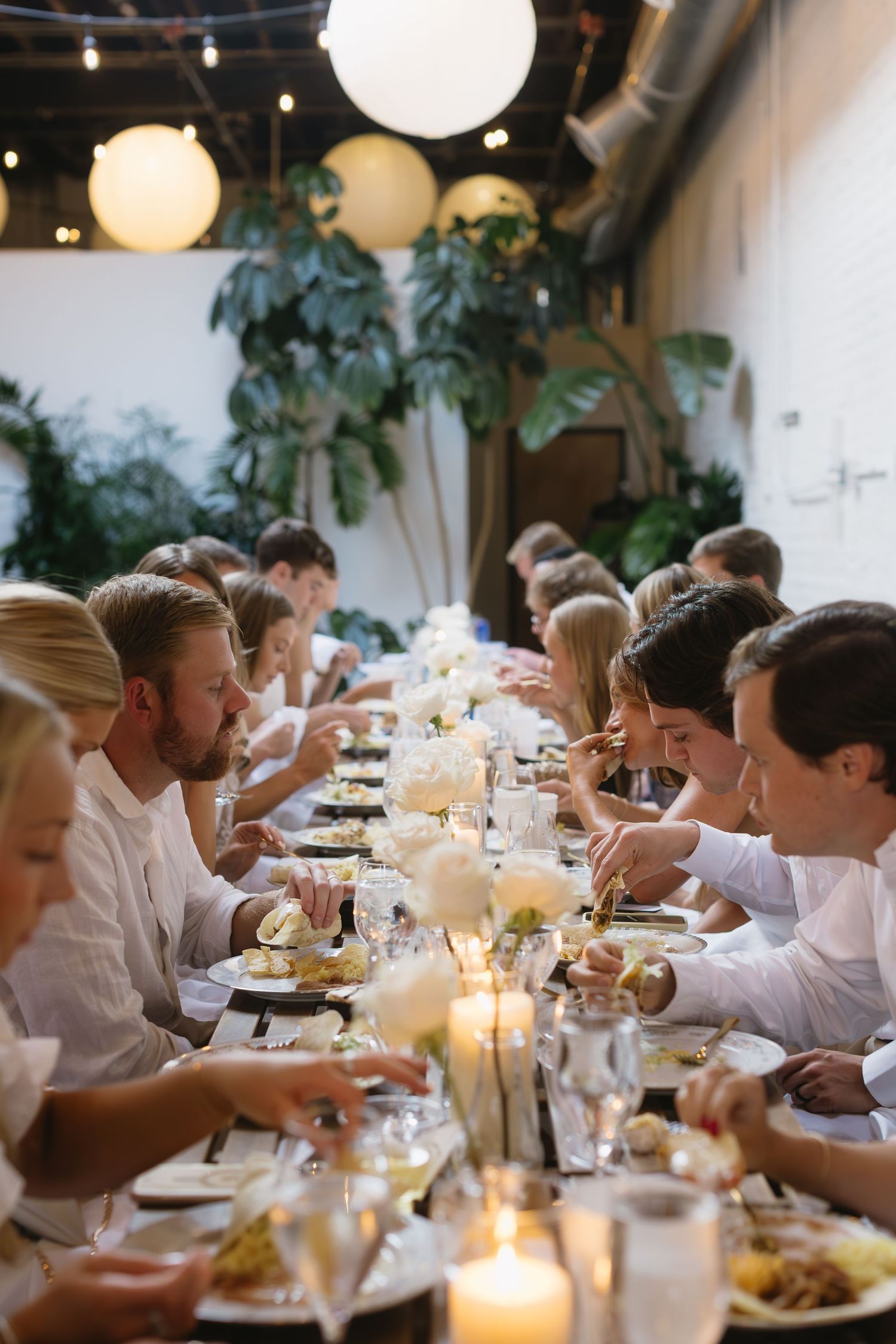 Candlelit dinner tables with guests in white clothing surrounded by indoor plants and paper lanterns.