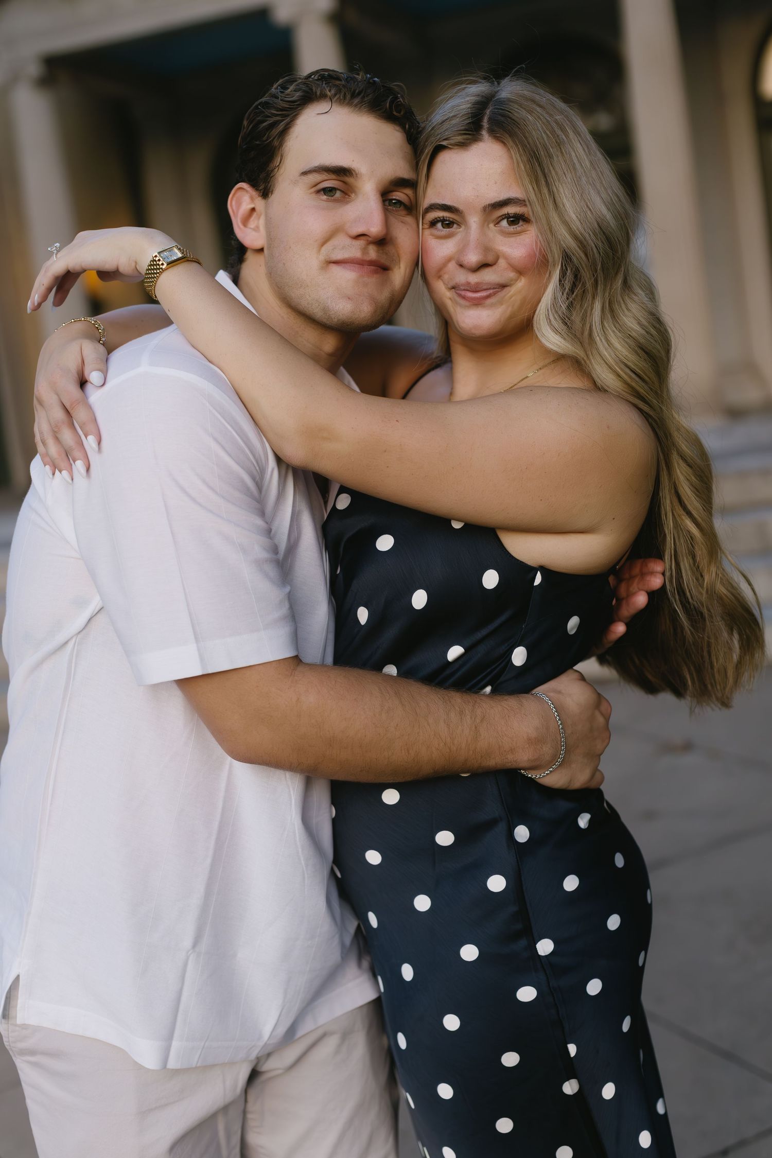 A couple embraces in a romantic moment wearing a white shirt and polka dot dress against a light architectural background.