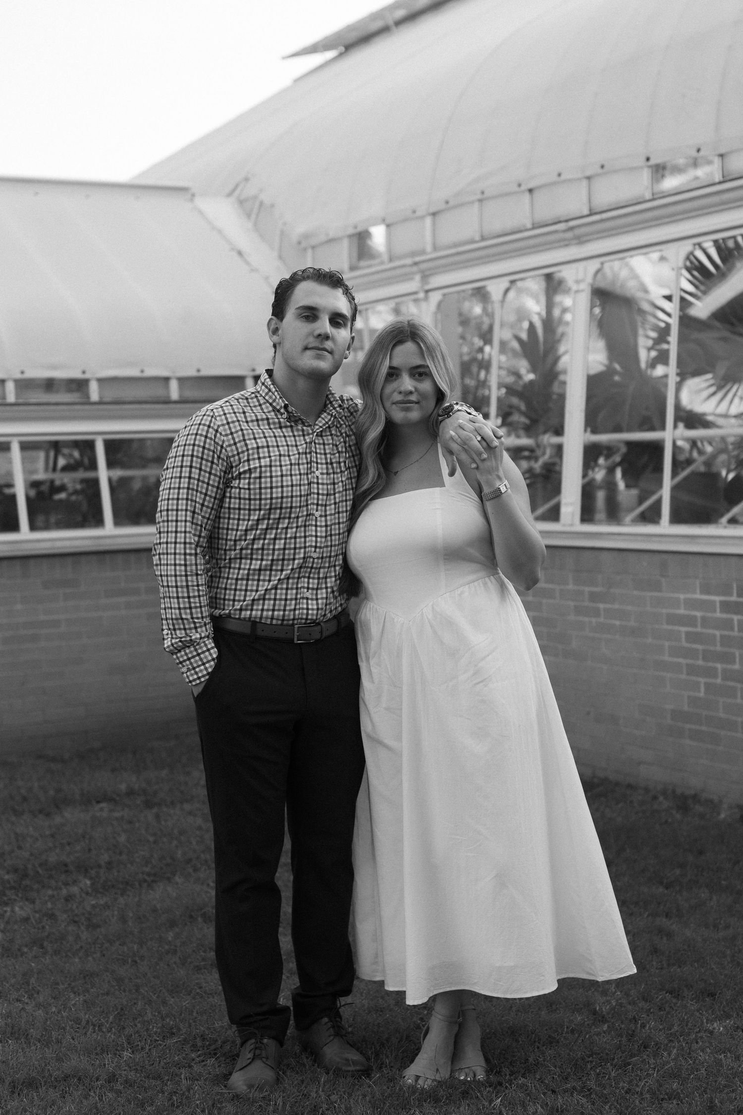 A couple stands together on grass in front of a brick building with windows, captured in black and white.