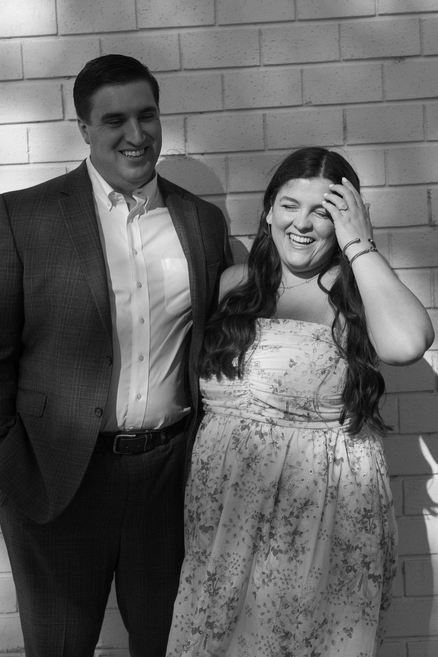 A couple laughs together during a romantic photoshoot against a white brick wall.