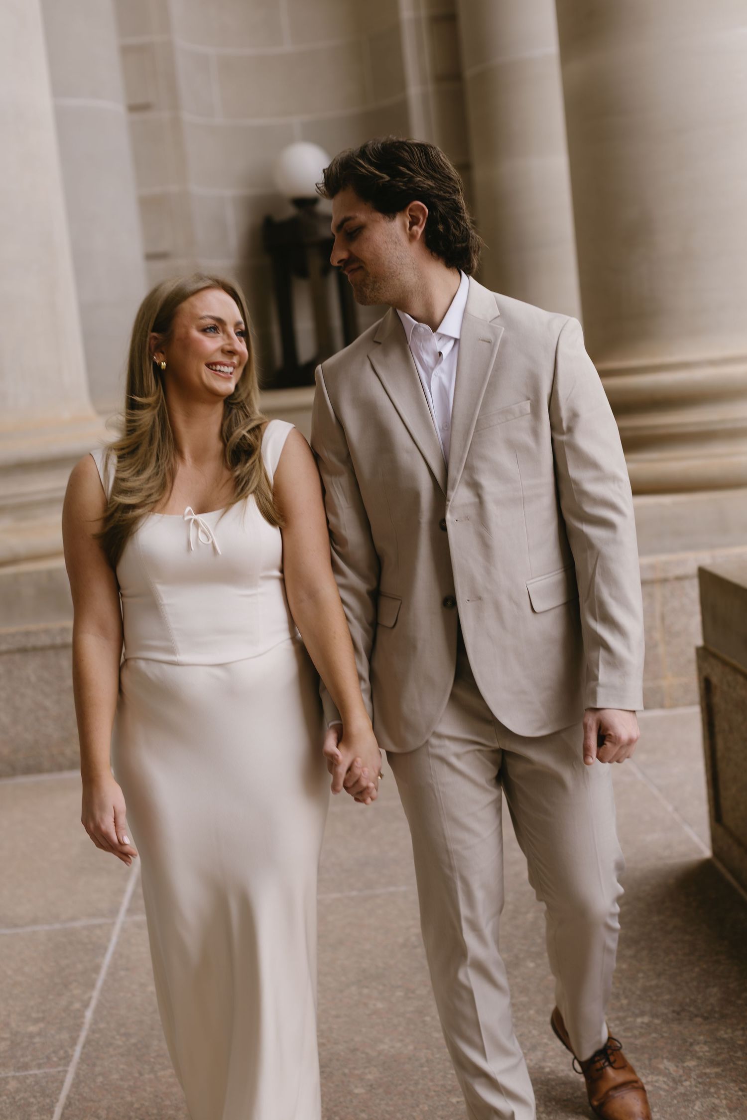 A couple in coordinated beige and white attire walks hand in hand along a columned walkway.