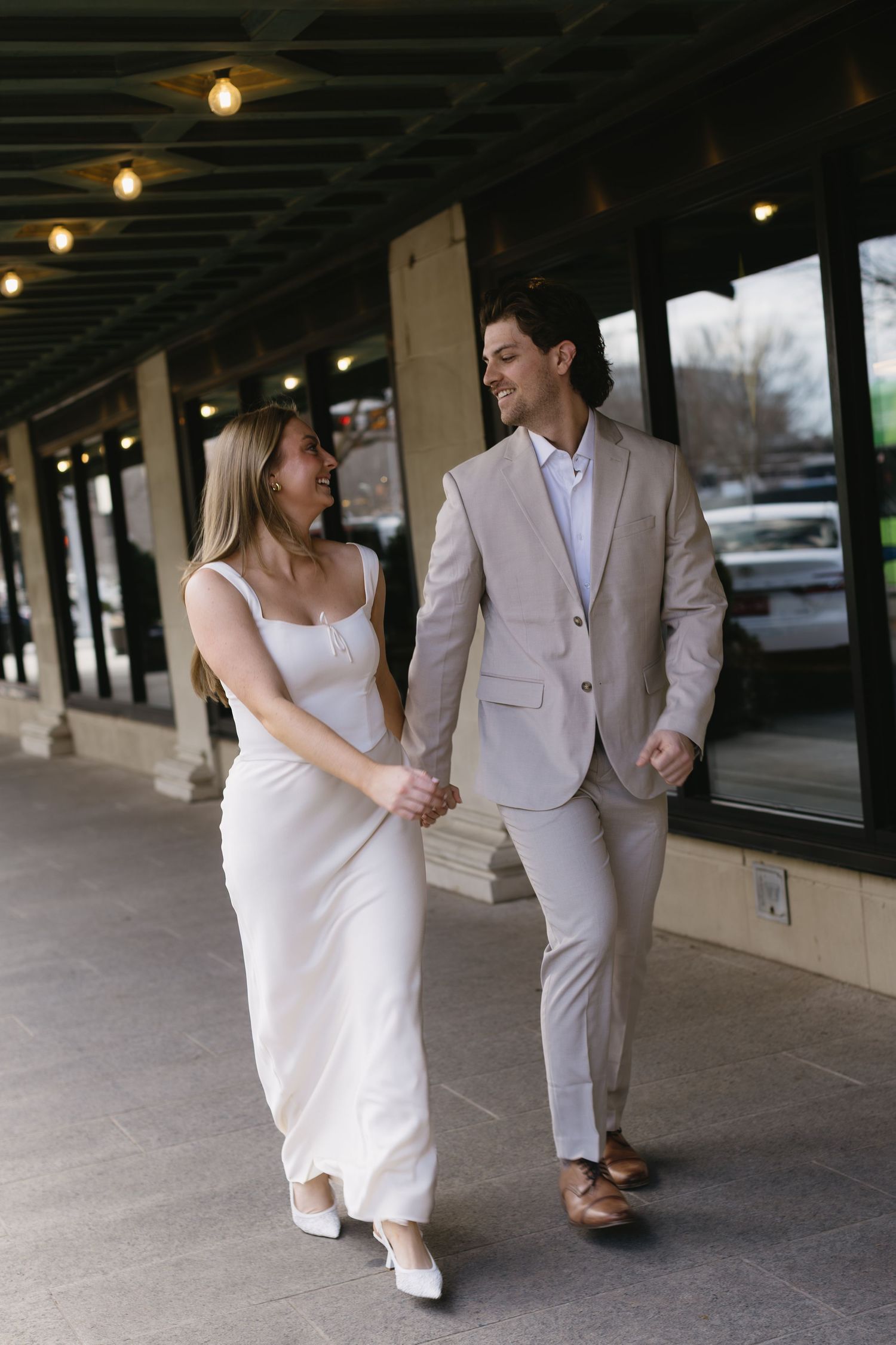 A couple walks hand in hand under covered walkway with string lights overhead.