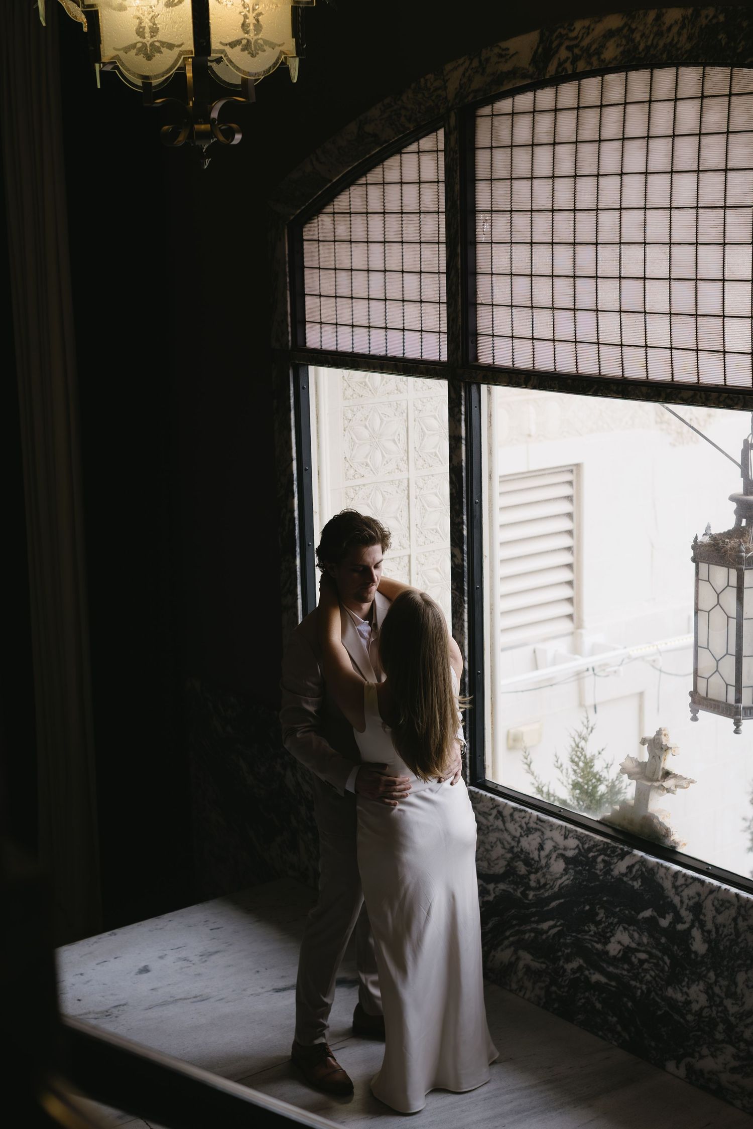 A couple shares an intimate moment in a dimly lit room with large windows.
