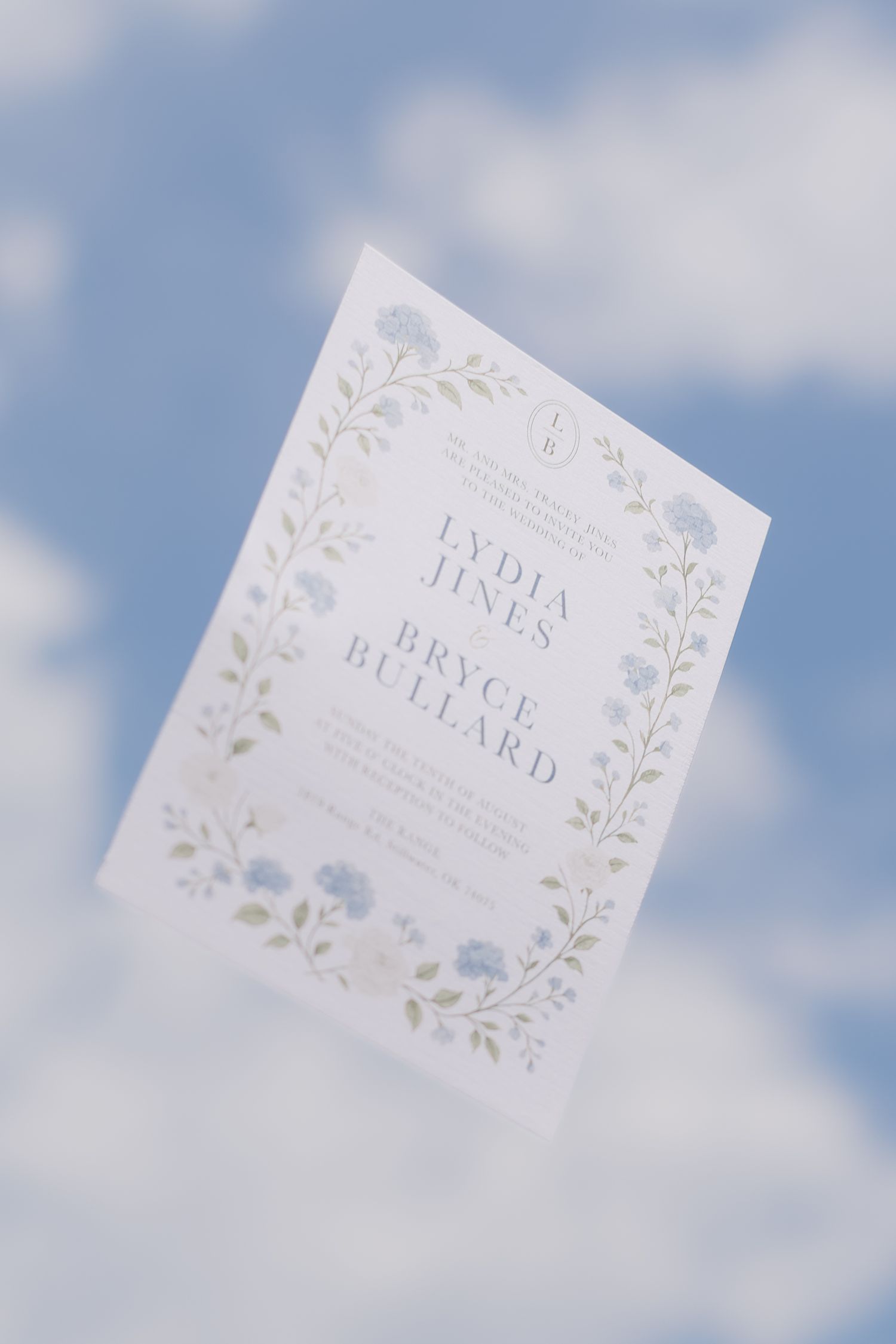 An elegant white wedding invitation with delicate floral details and soft blue flowers photographed against a cloudy sky.