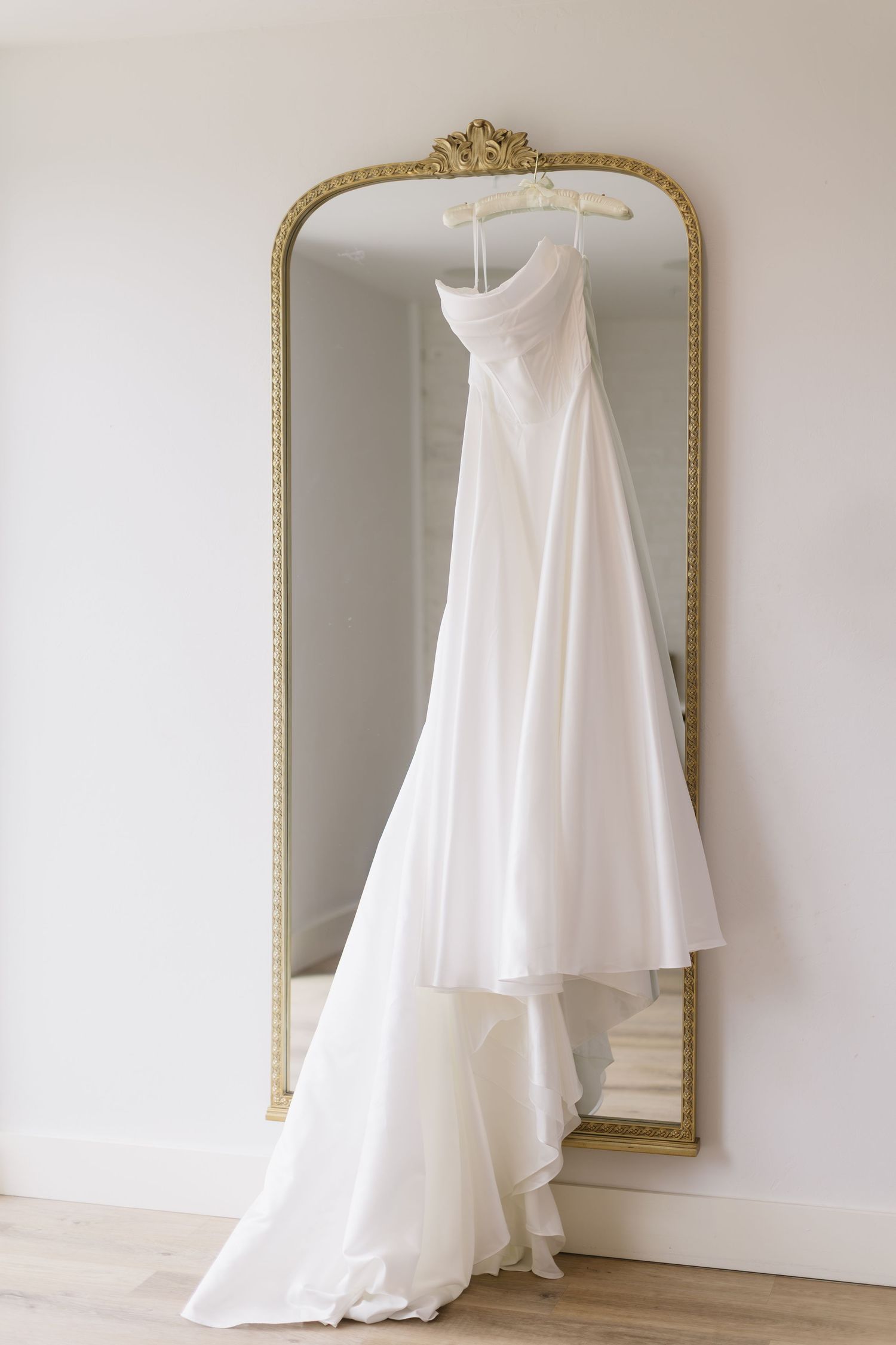 An elegant white wedding dress hangs on display against a decorative gold-framed mirror.