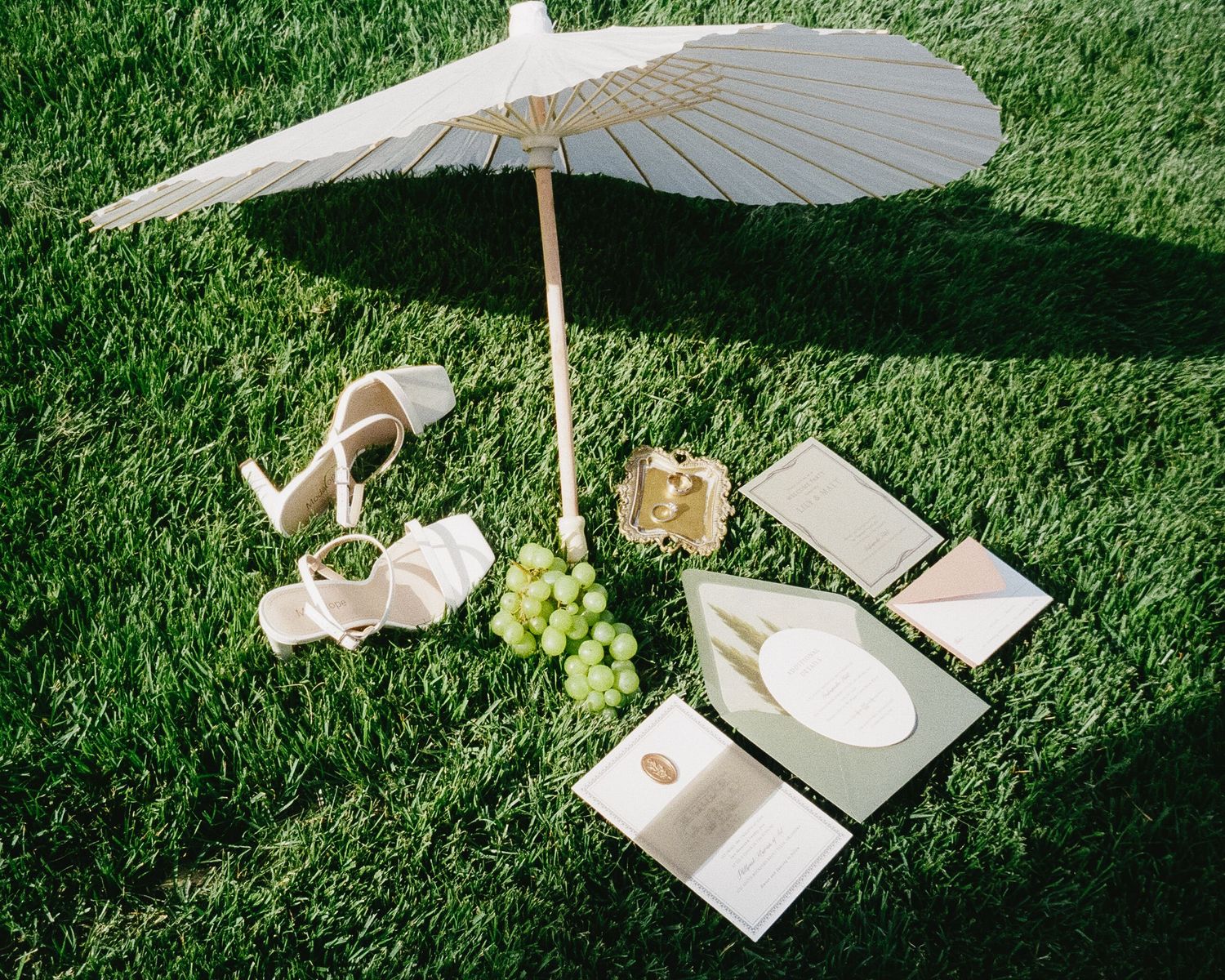 A white parasol shades a picnic setup with champagne, grapes, and elegant place settings on green grass.