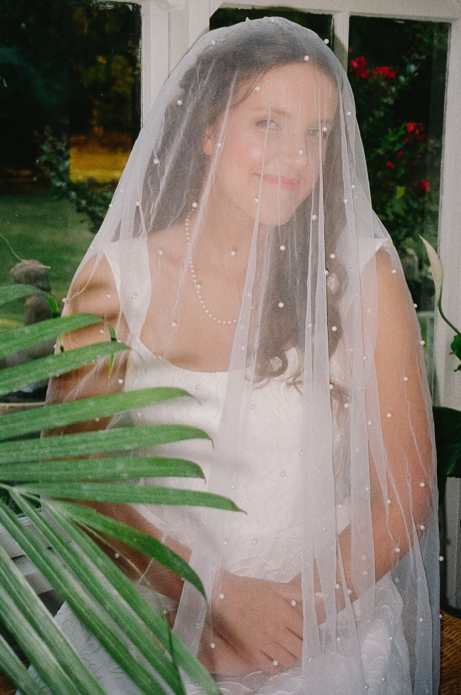 Ethereal bridal portrait through a delicate pearl-studded veil with tropical foliage in foreground.