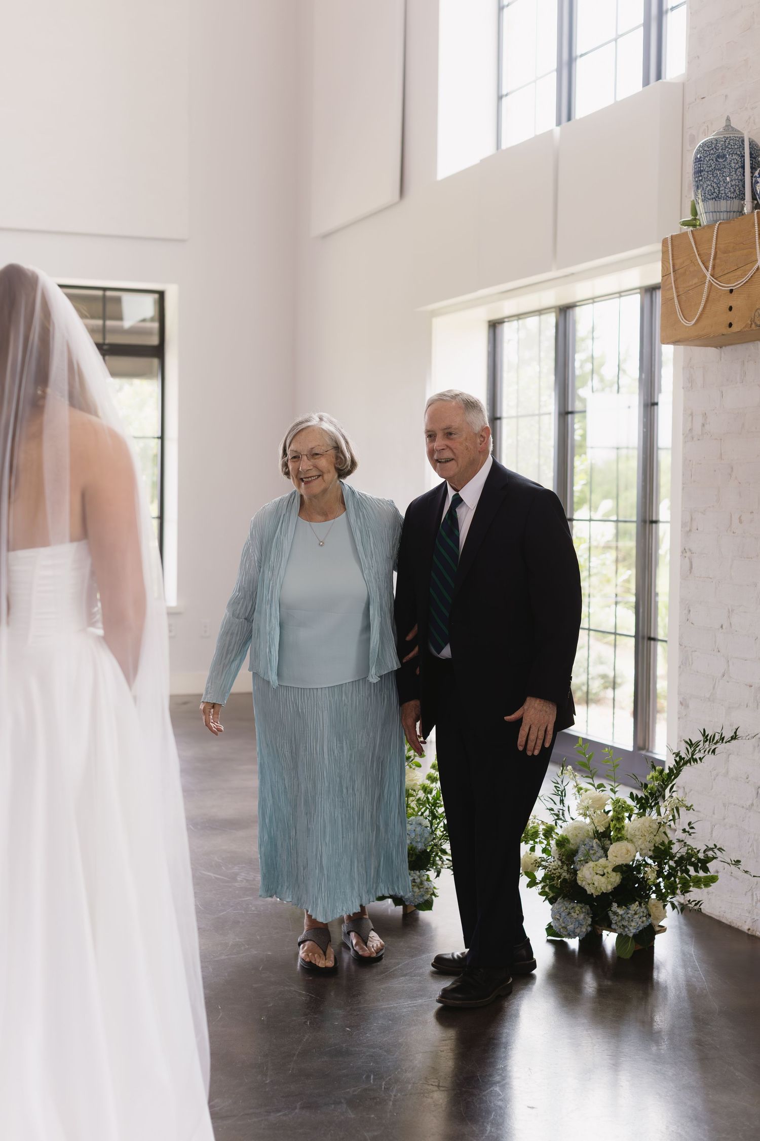 Wedding party members gathering in bright indoor venue with large windows.