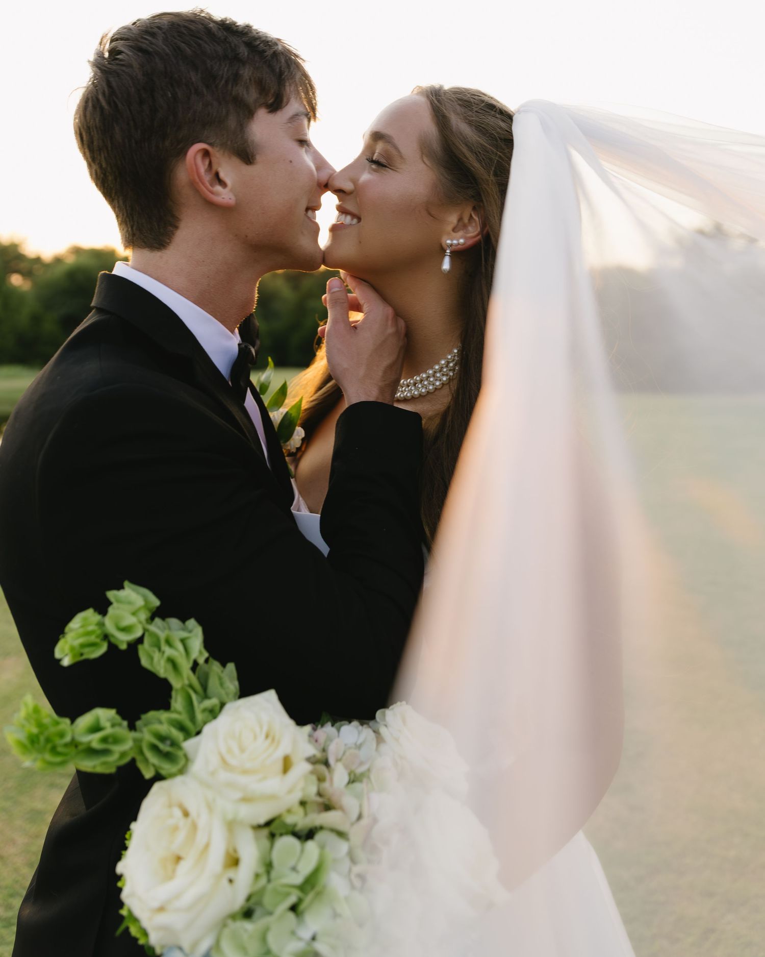 Romantic bride and groom share intimate moments at sunset with white bouquet against pastoral backdrop.