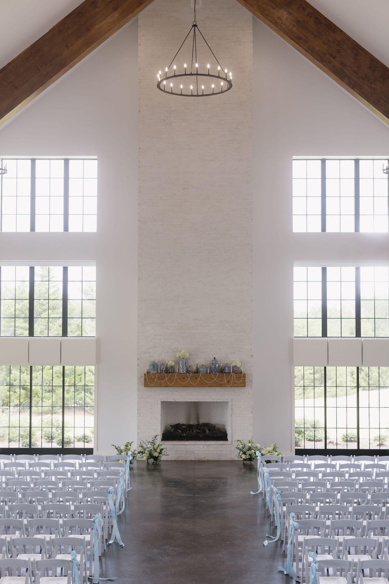 Modern white wedding ceremony space with exposed beams, large windows and rows of white chairs in a minimalist setting.