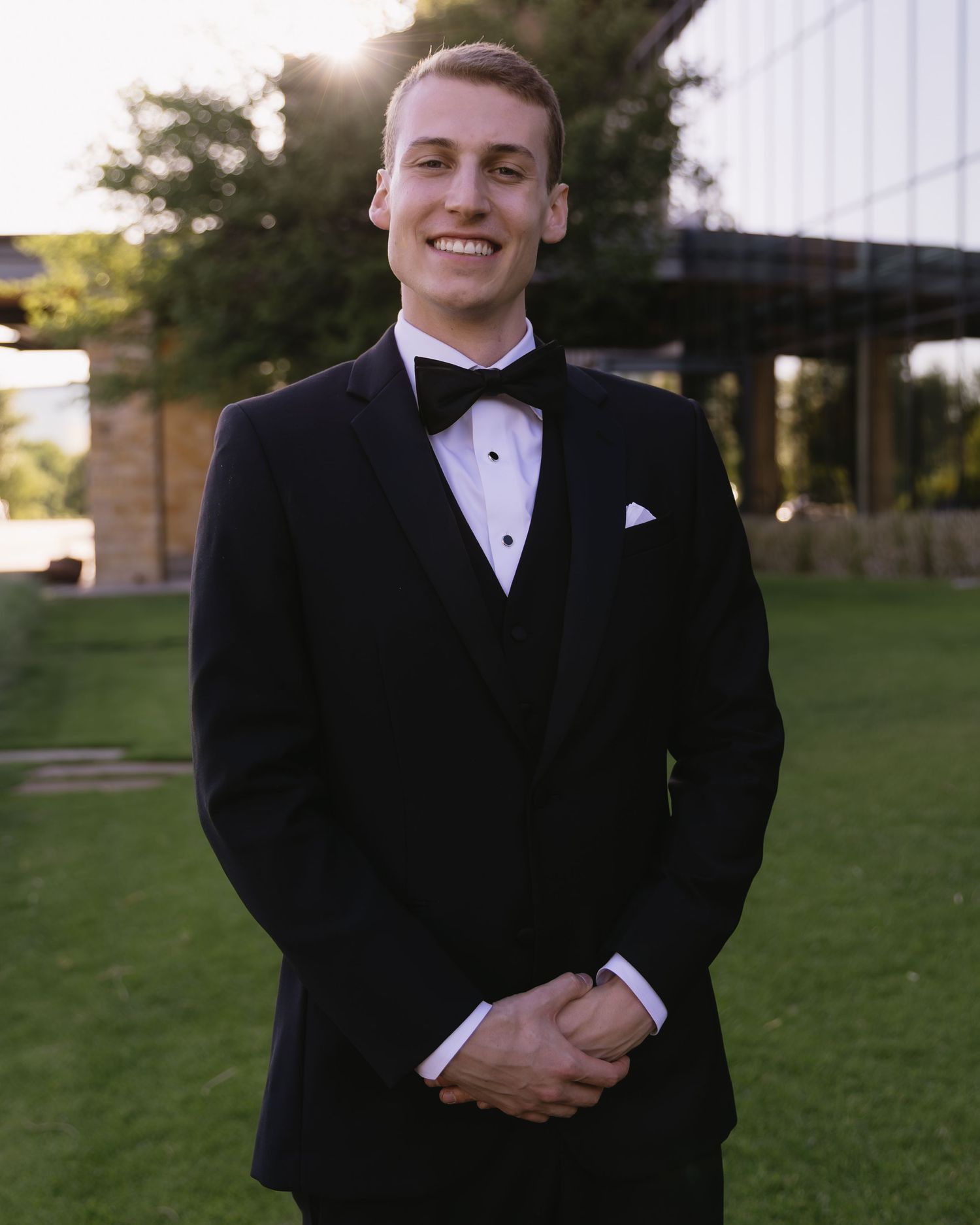 Groom in classic black tuxedo and bowtie standing on lawn with building in background.